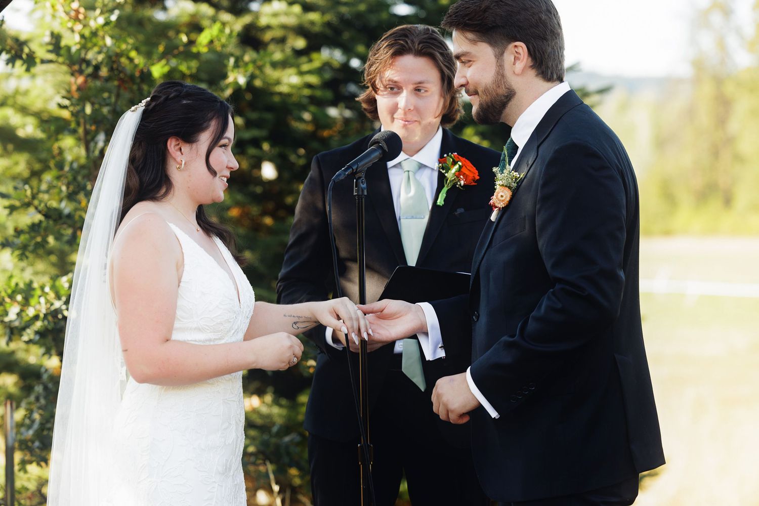 Bride and groom exchange rings during outdoor wedding ceremony with officiant standing behind them.