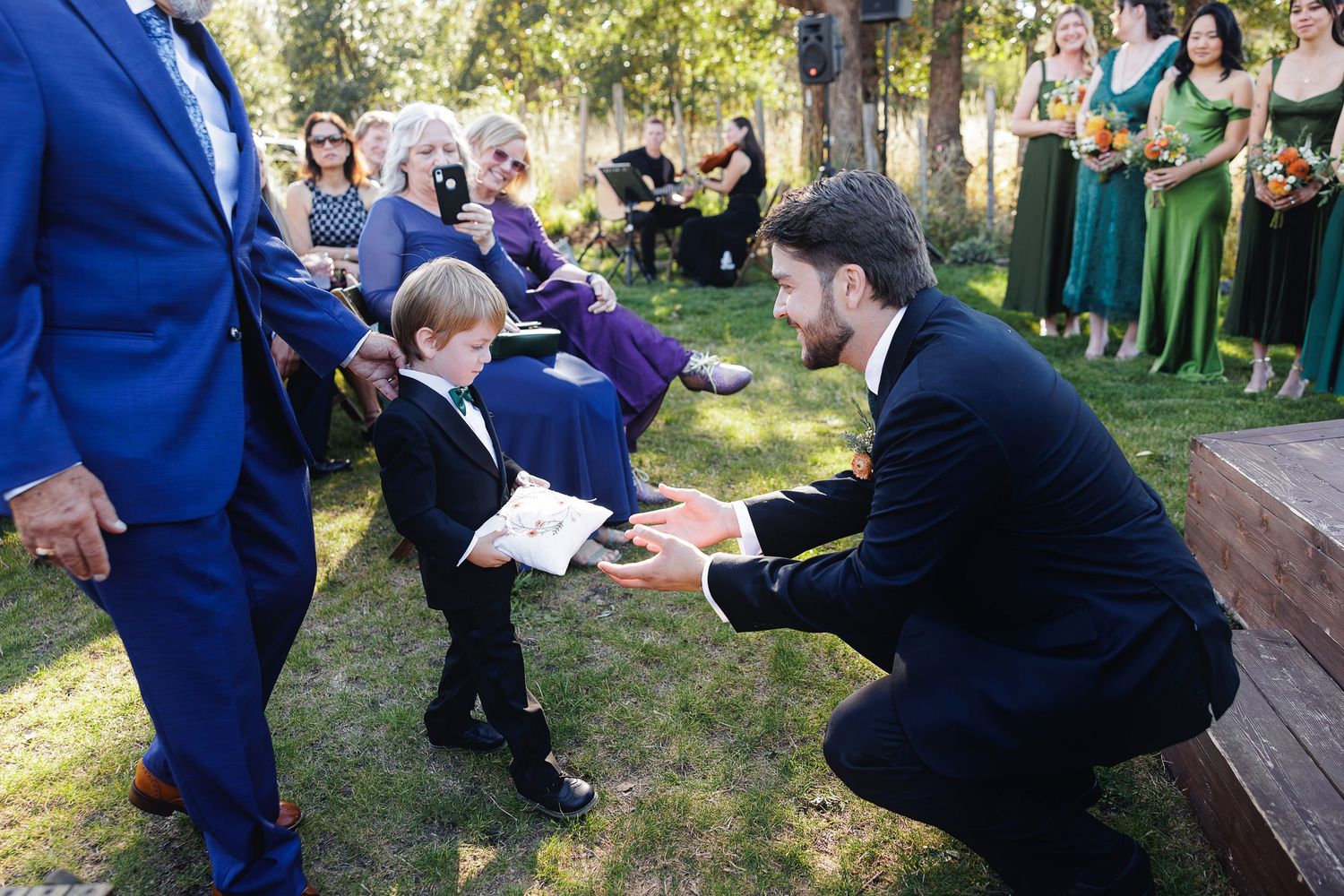 Ring bearer presenting wedding rings to groomsman during outdoor wedding ceremony with guests and bridesmaids in background.