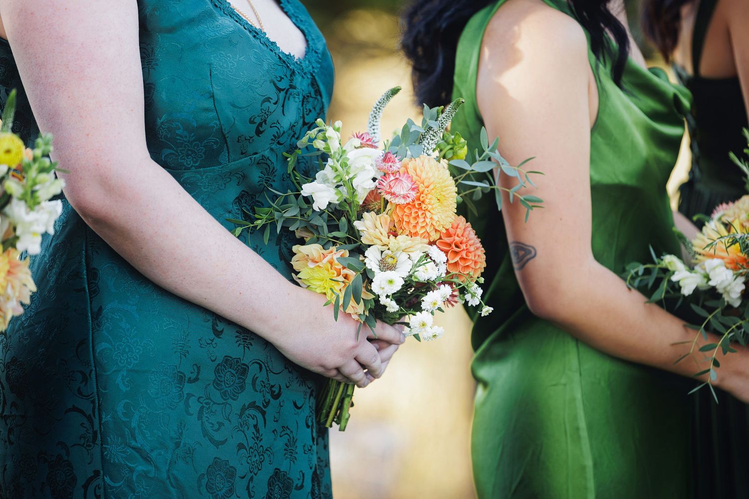 Two bridesmaids in teal and green dresses holding colorful wildflower bouquets with orange dahlias and white blooms.