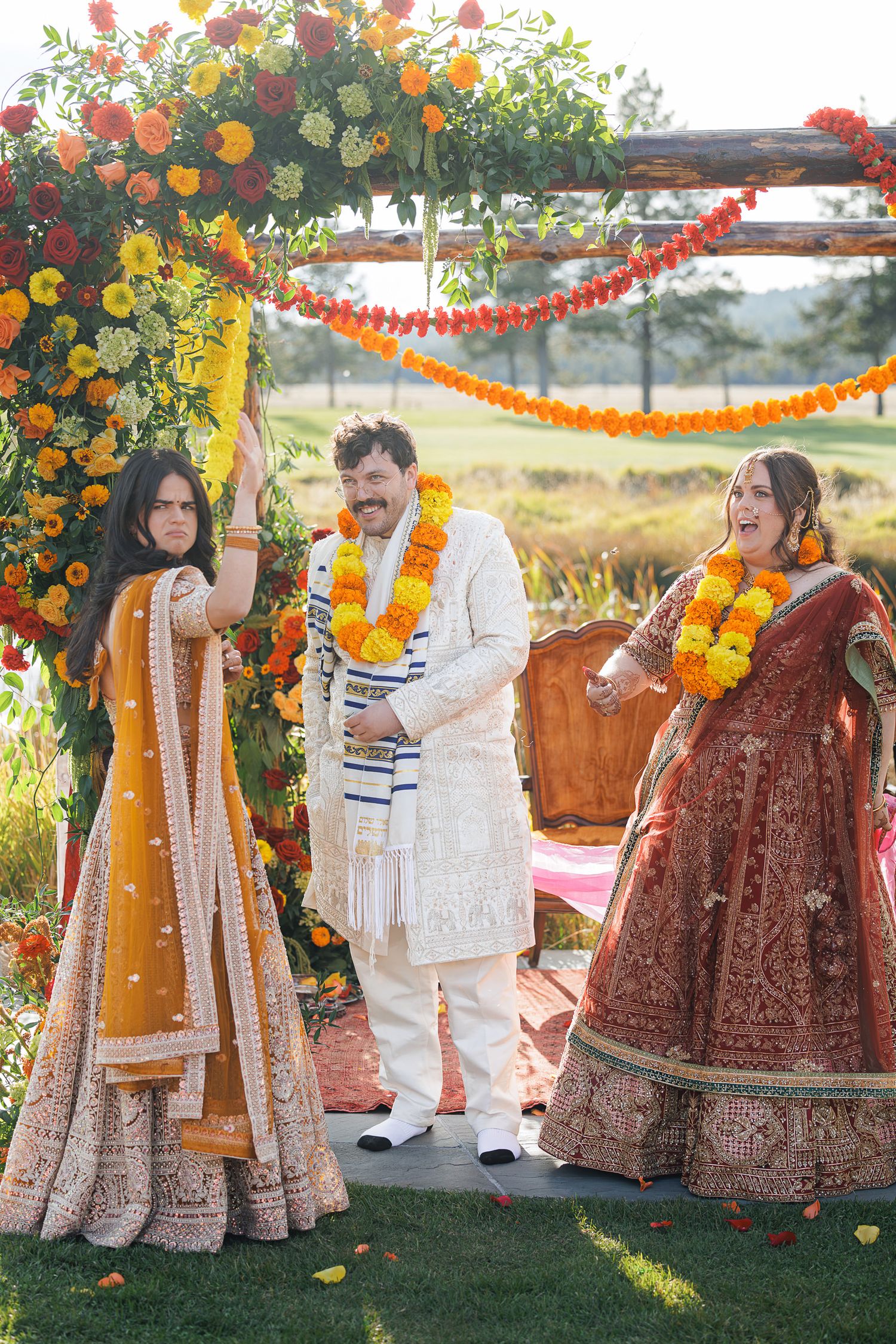 Joyful Indian wedding ceremony with bride in red lehenga, groom in white sherwani, and bridesmaid in yellow lehenga under floral mandap.