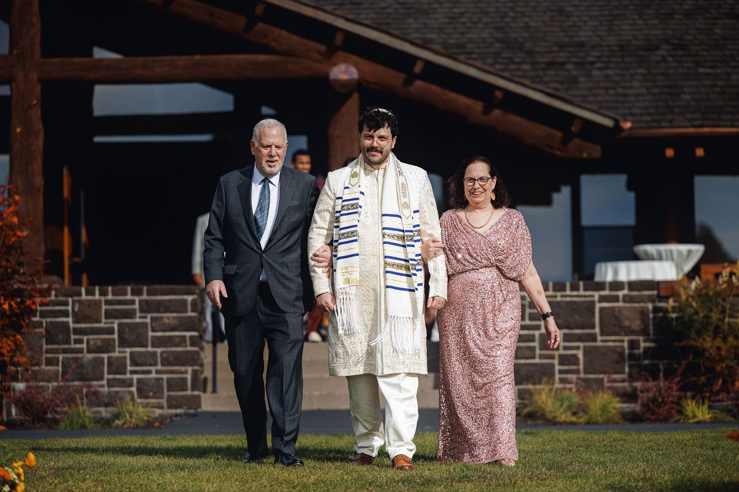 Three people in formal attire walk together outdoors, with the center person wearing a tallit at a Jewish celebration.