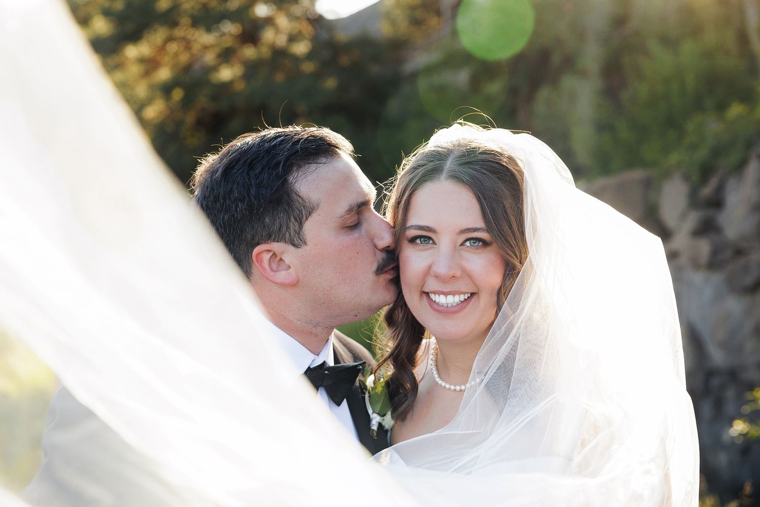 Groom in black tuxedo kissing bride on cheek, bride smiling in white gown and veil on wedding day.