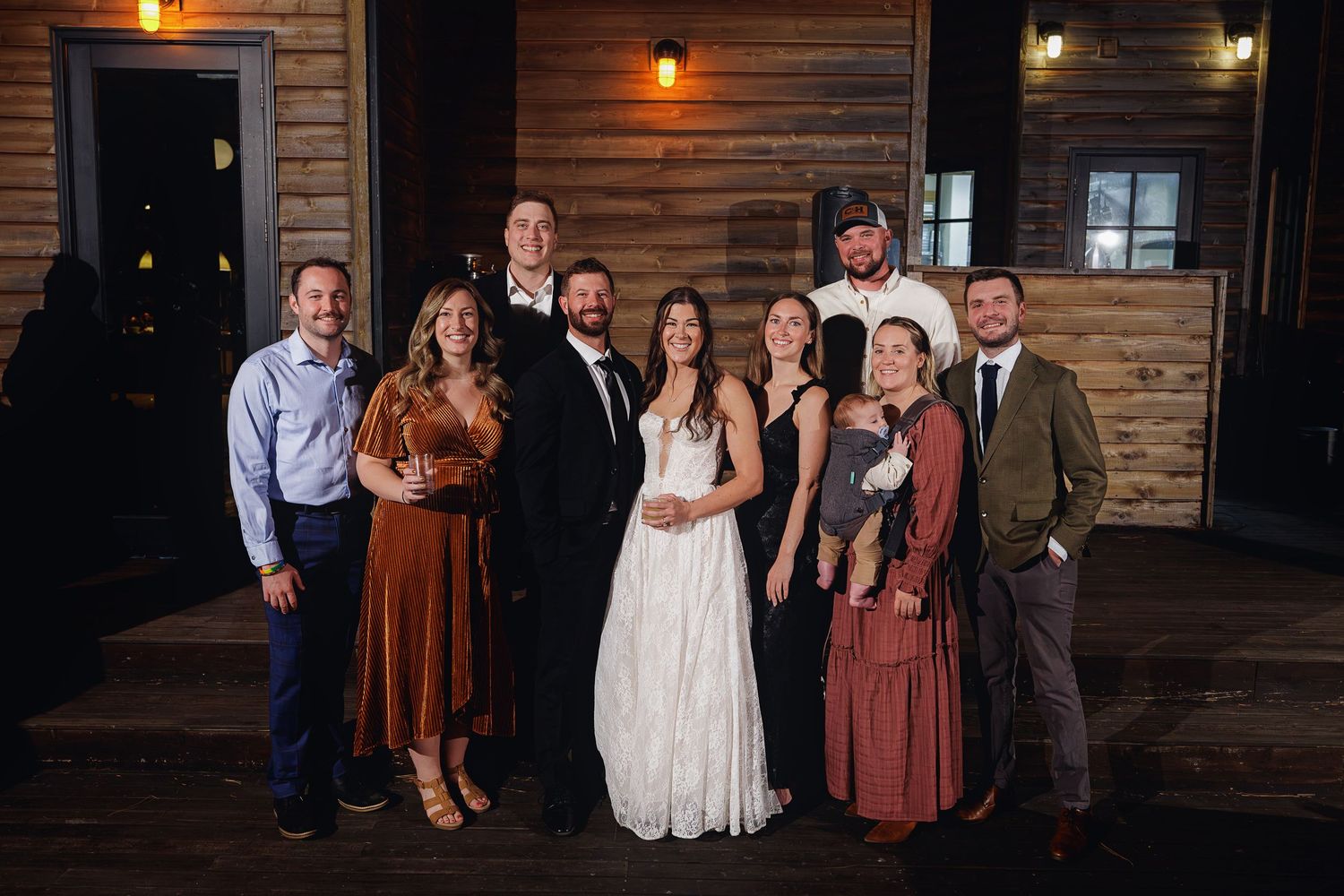 Wedding party poses together in front of a rustic wooden venue, bride in white gown surrounded by family and friends.