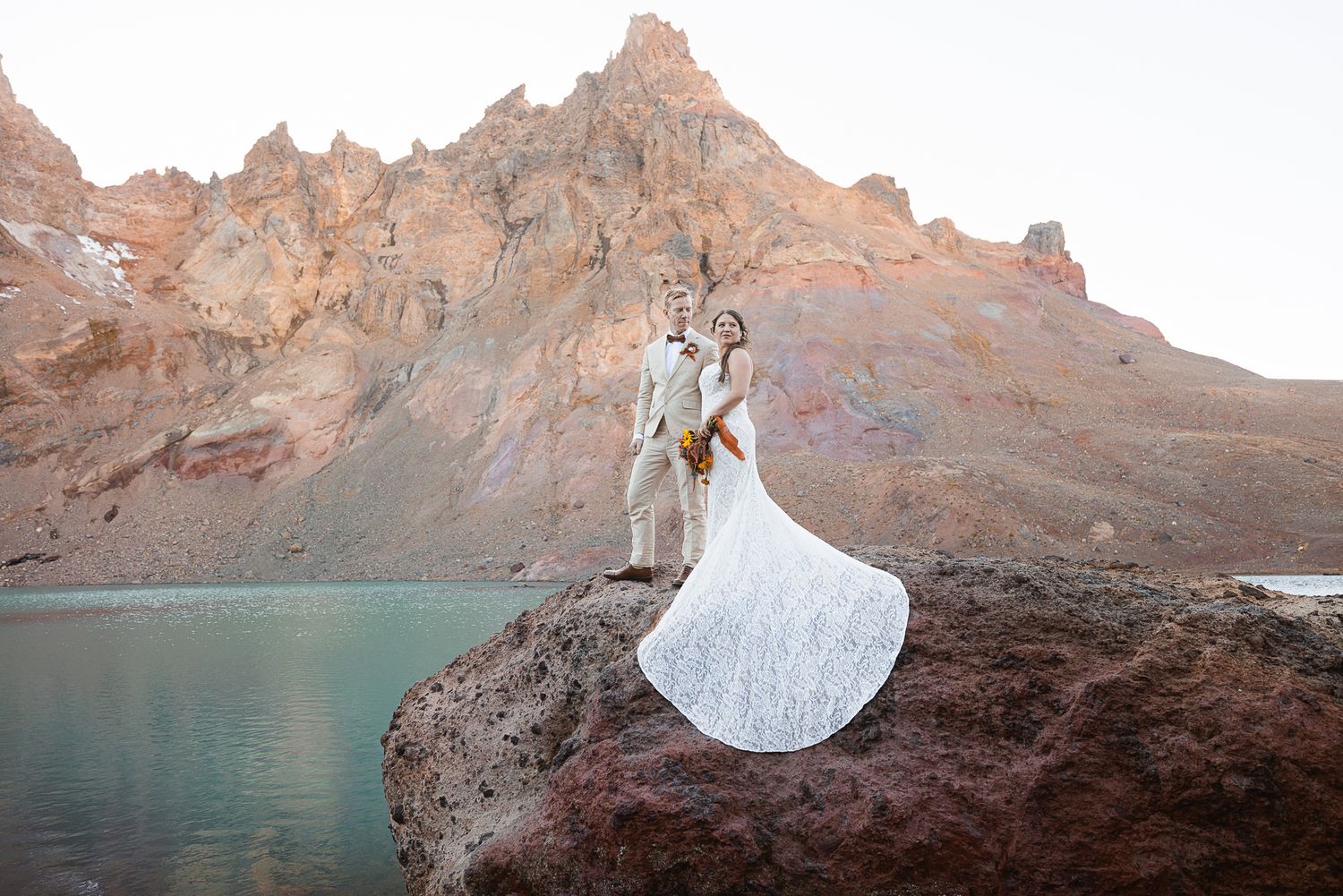 Bride in a flowing white gown and groom in a light suit embrace on a rock beside a turquoise lake with rugged orange mountains behind.