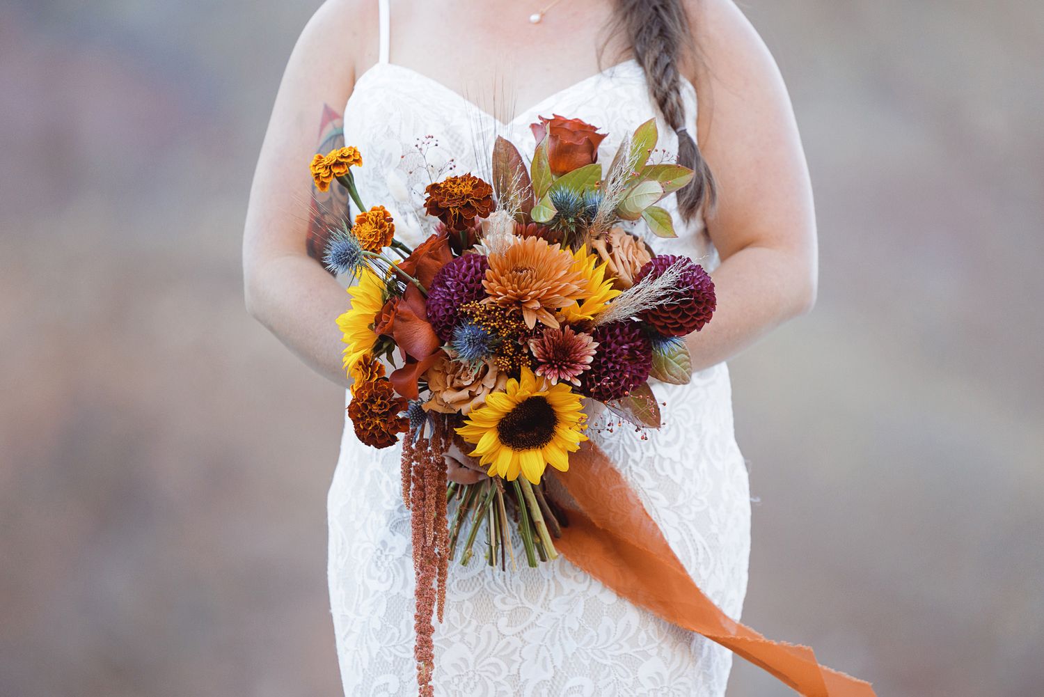 Bride in a white lace wedding dress holding an autumn bouquet of sunflowers, orange blooms and burgundy flowers with a rust ribbon.