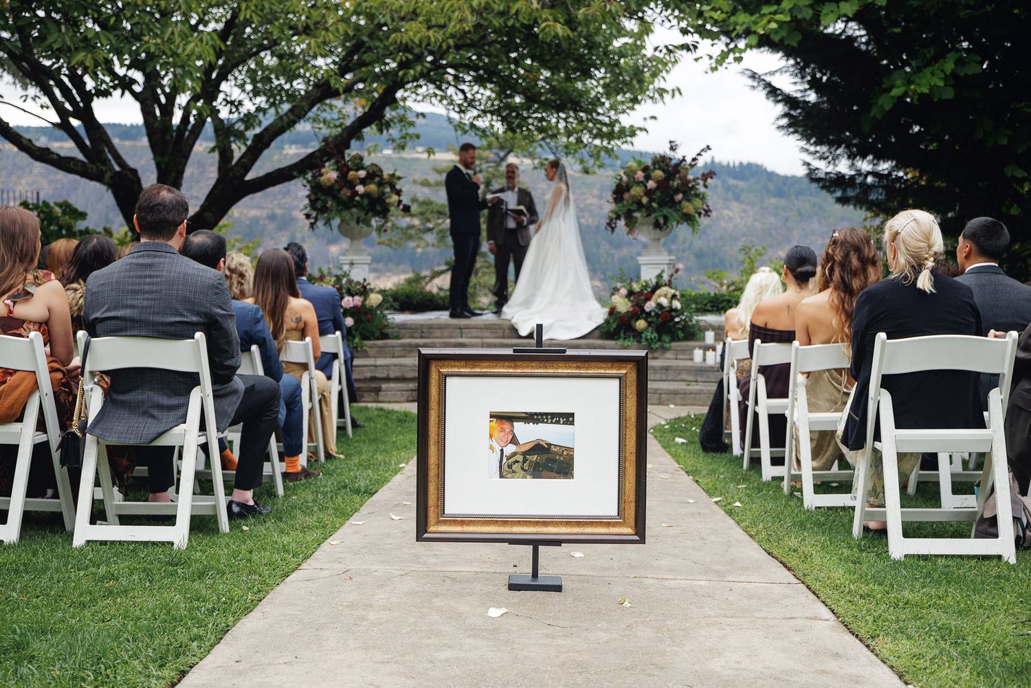 Outdoor wedding ceremony with guests seated on white chairs, couple at altar, framed photo display on aisle.