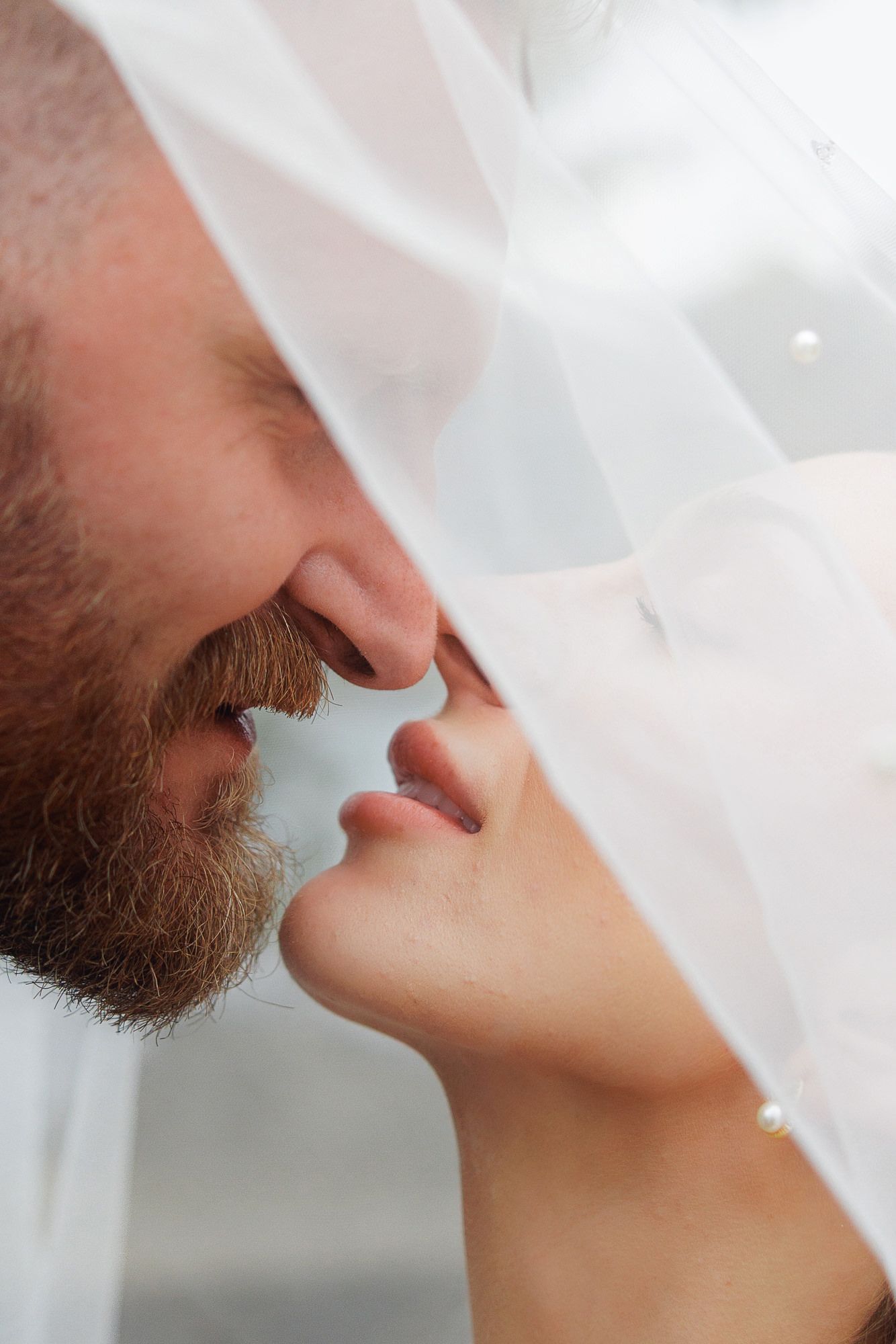 Bride and groom sharing a romantic moment under a white wedding veil, close-up of their faces nearly kissing.