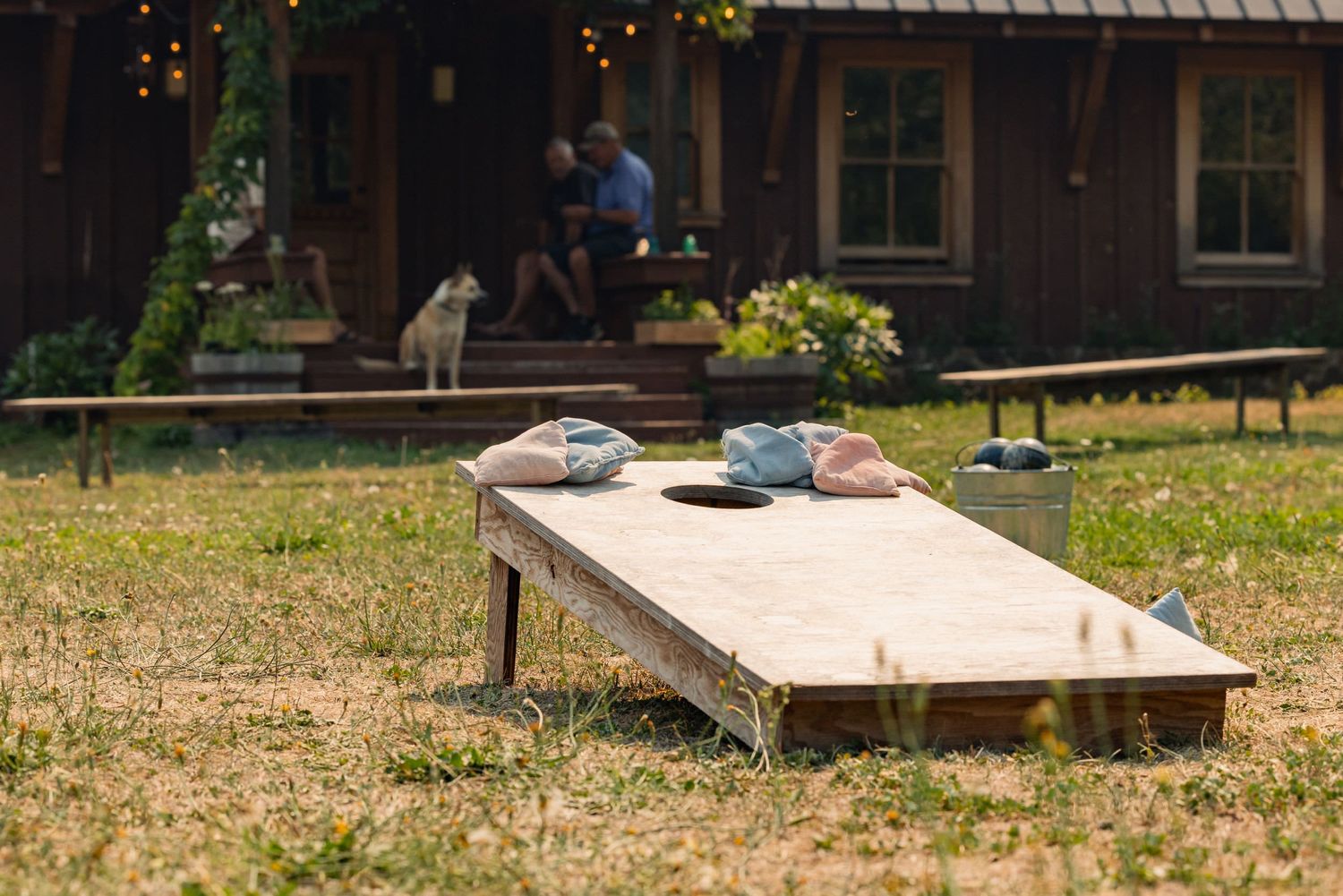 A cornhole board sits on grassy ground with a rustic cabin and picnic tables in the background.
