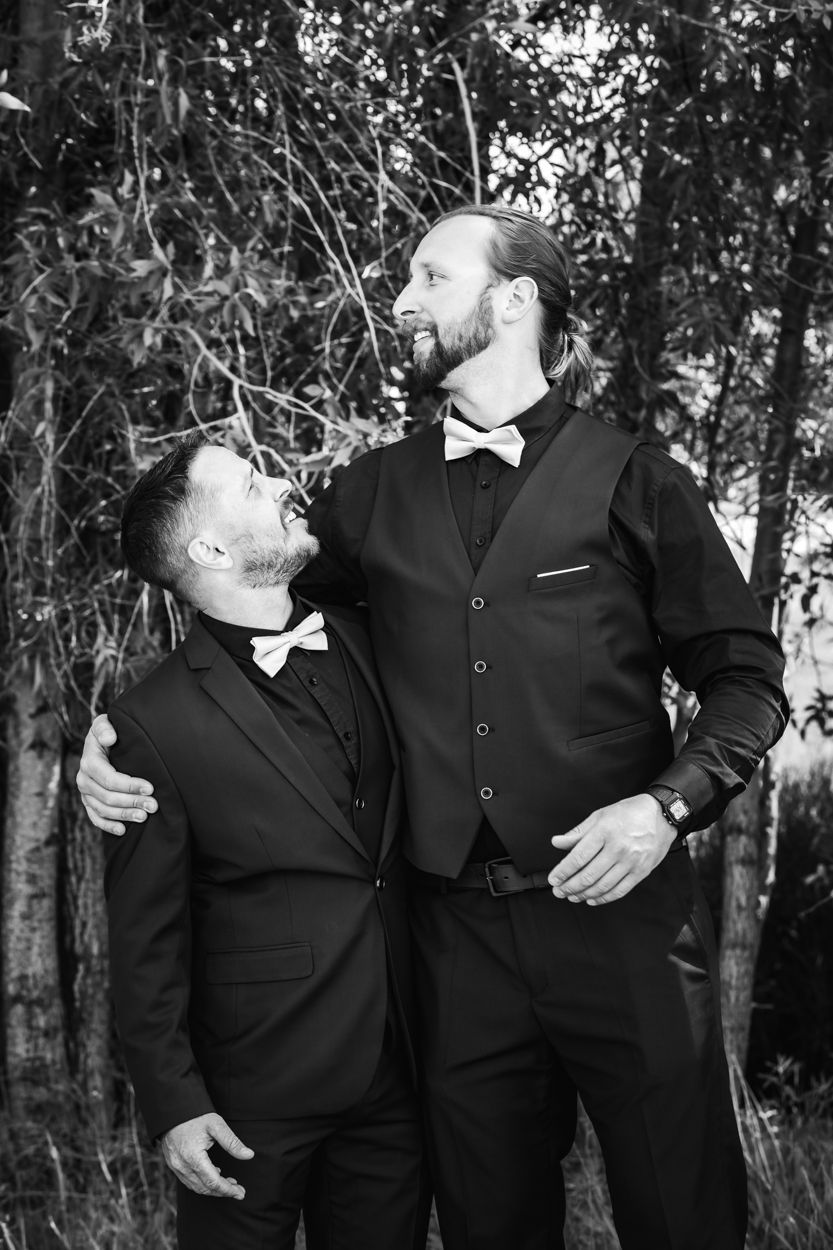 Two people wearing black formal attire and bow ties share a joyful moment together outdoors in a black and white photo.