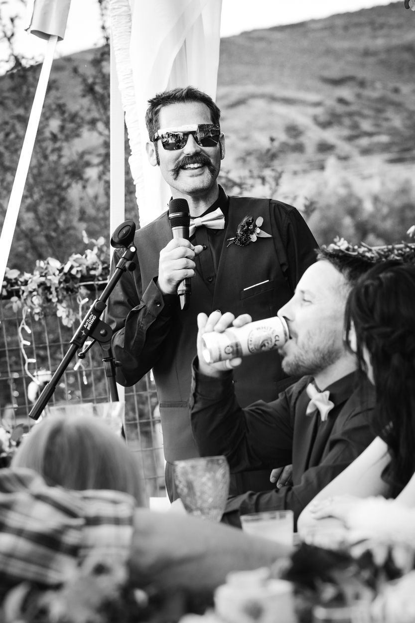 A speaker in sunglasses and a dark suit holds a microphone while giving a speech at an outdoor event with tables and guests.