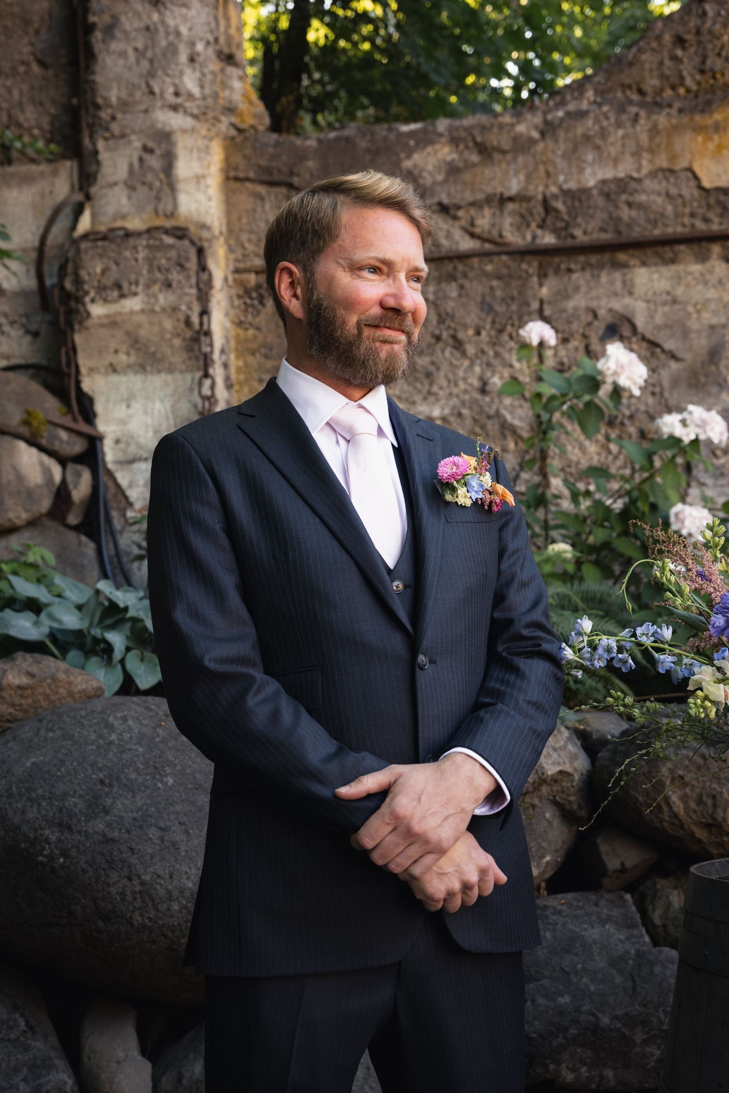 Groom wearing a navy three-piece suit with pink boutonniere stands in front of stone wall with blooming flowers.