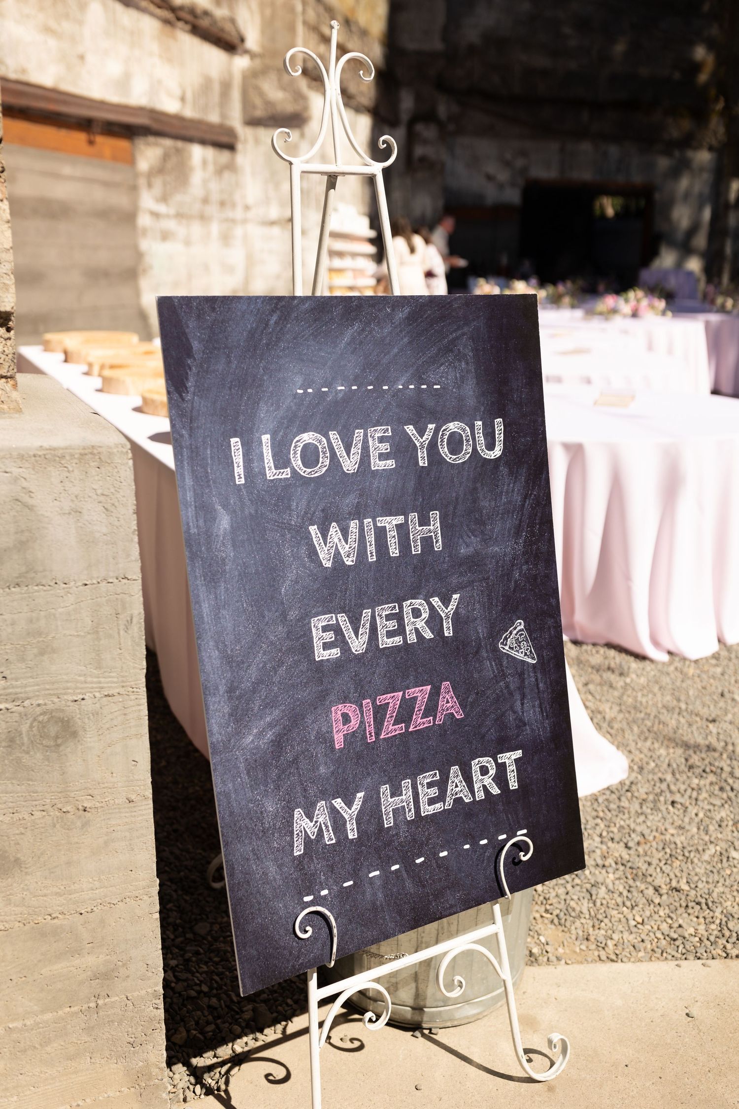 A chalkboard sign with cursive writing reads 'I Love You With Every Pizza My Heart' displayed on a decorative metal stand.