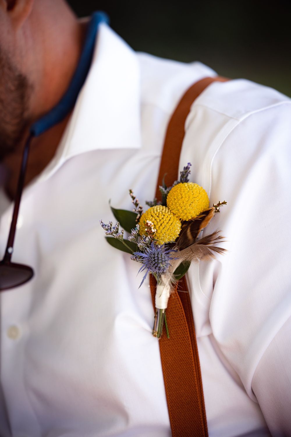 Wedding boutonniere with yellow craspedia flowers, blue thistle, and greenery pinned to brown suspenders and white shirt.