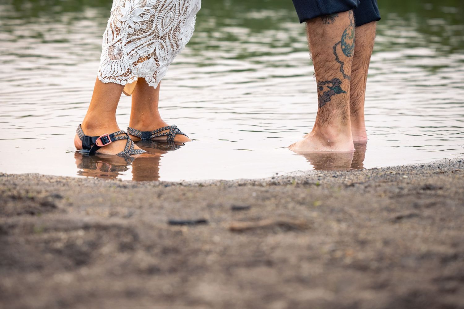Two pairs of bare feet standing in shallow water at a beach shore, one with visible leg tattoos.