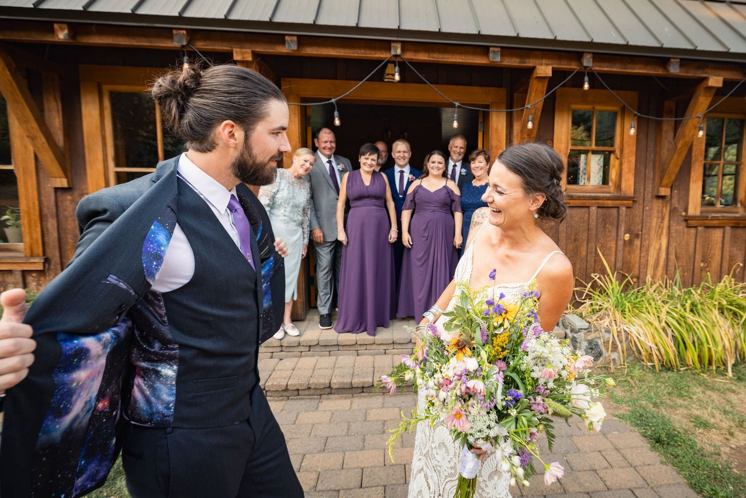 A joyful wedding moment captured outside a rustic wooden venue with bridesmaids in purple dresses looking on.