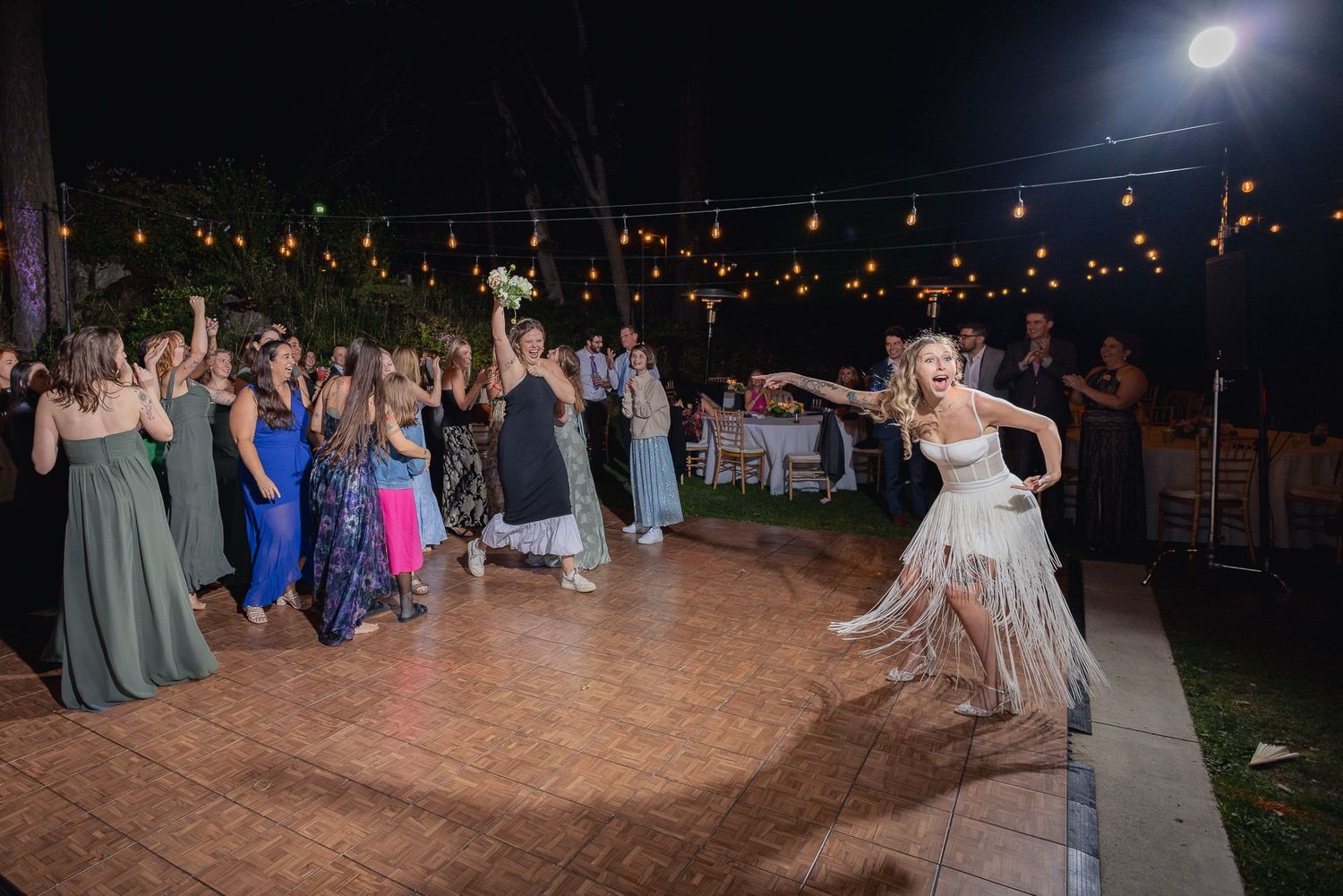 A festive outdoor wedding reception dance party at night with string lights overhead.