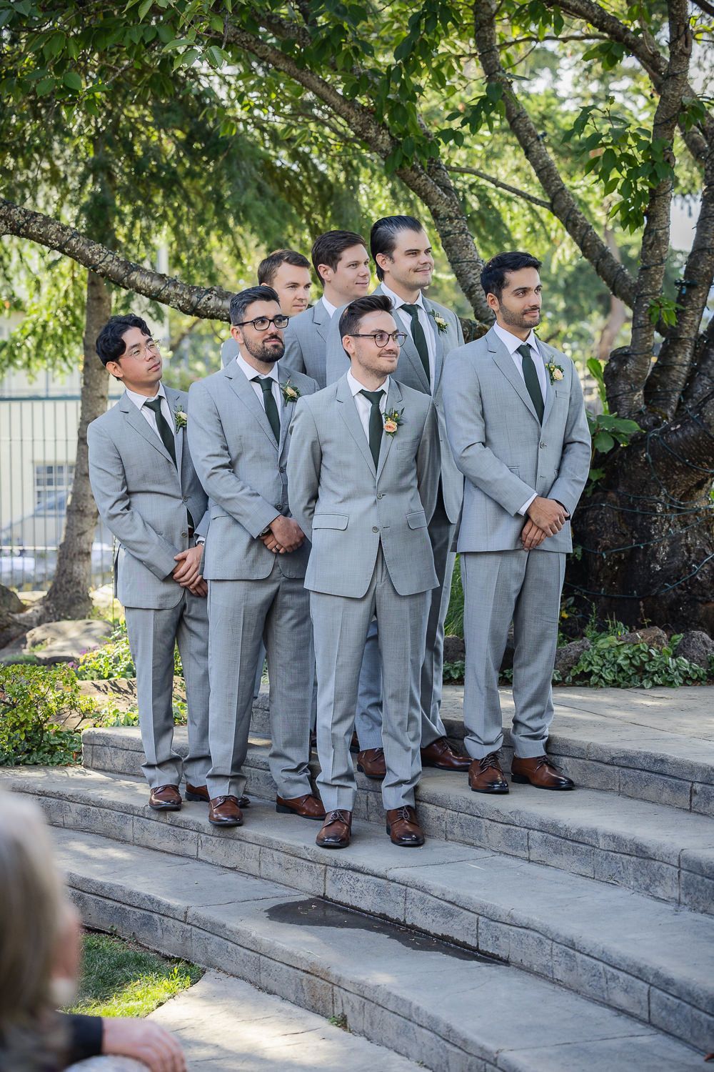 A group of groomsmen in matching light gray suits stand together on outdoor stone steps under a leafy green tree.