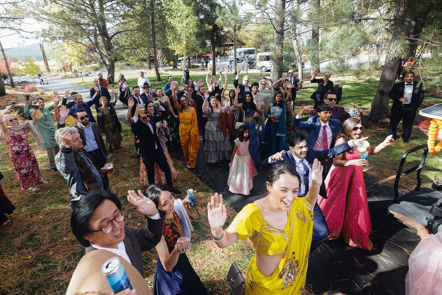 Large group of guests dancing outdoors at a colorful multicultural wedding celebration in a park setting.