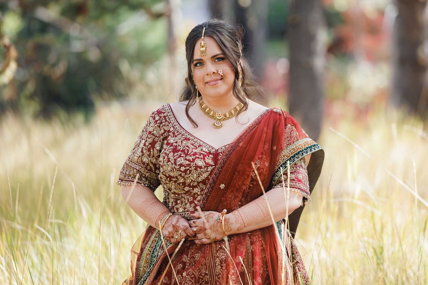 Woman in red embroidered saree with gold jewelry poses gracefully in a sunlit field of tall golden grass.