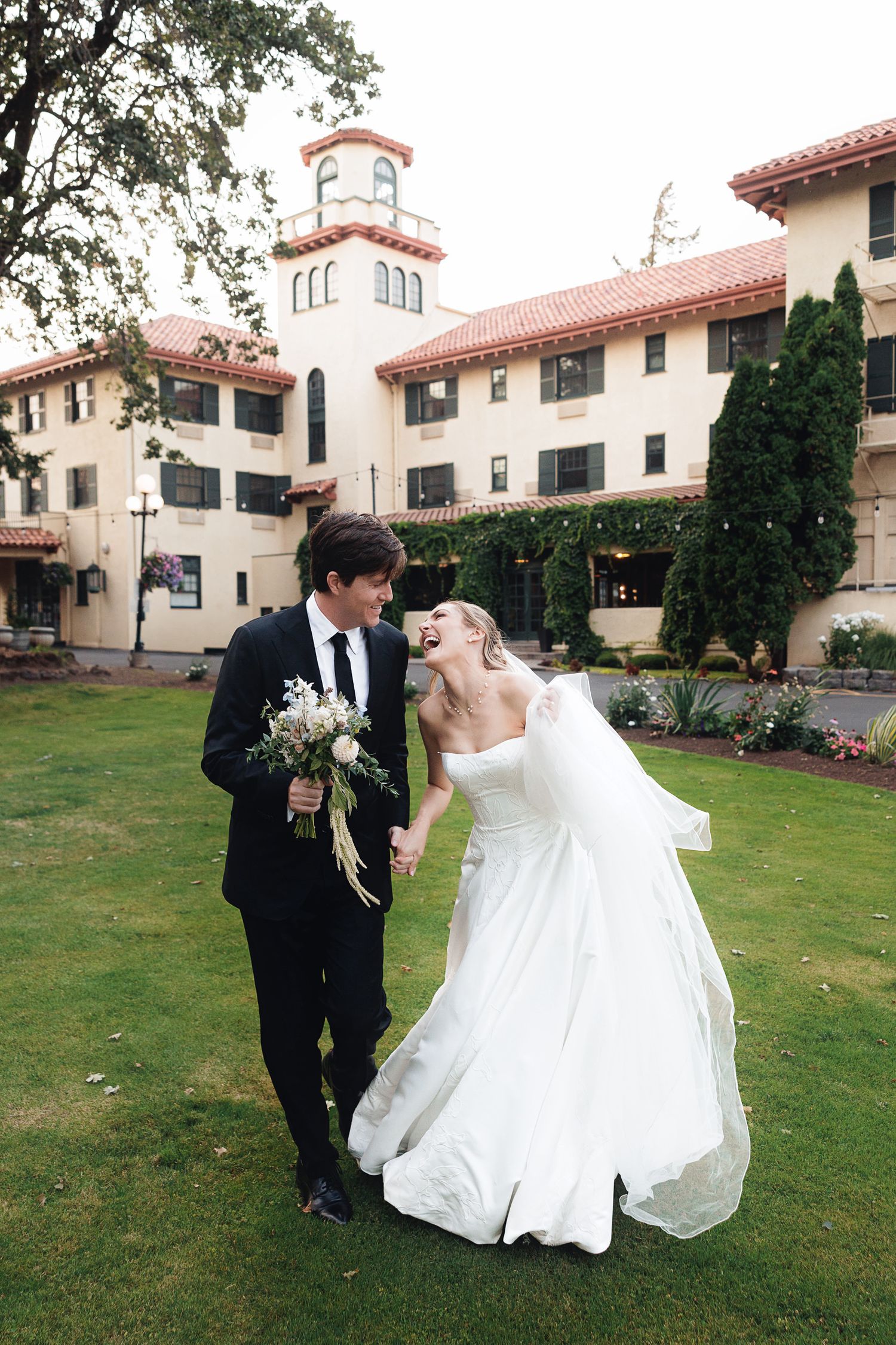 Bride and groom laughing together on green lawn outside elegant Spanish-style wedding venue.