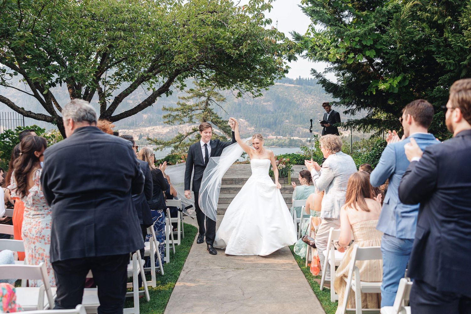Bride and groom celebrate walking down the aisle at an outdoor wedding ceremony with guests applauding.