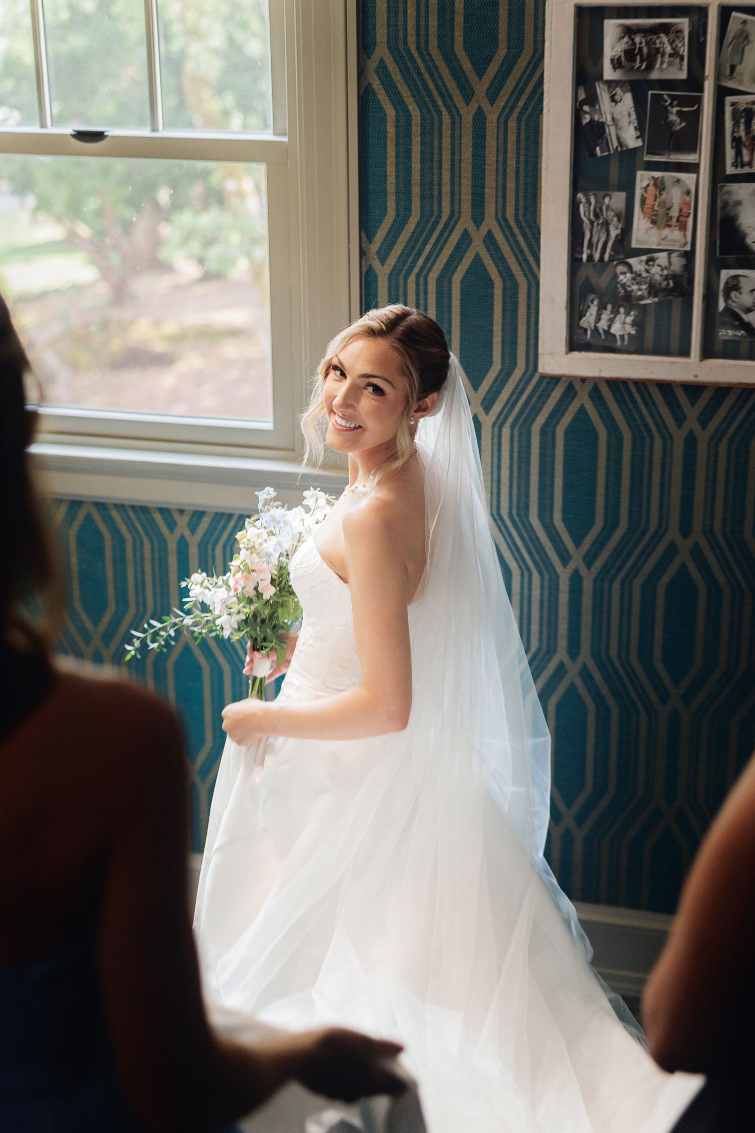 Bride in white gown and veil holds bouquet, smiling by window with teal geometric wallpaper backdrop.