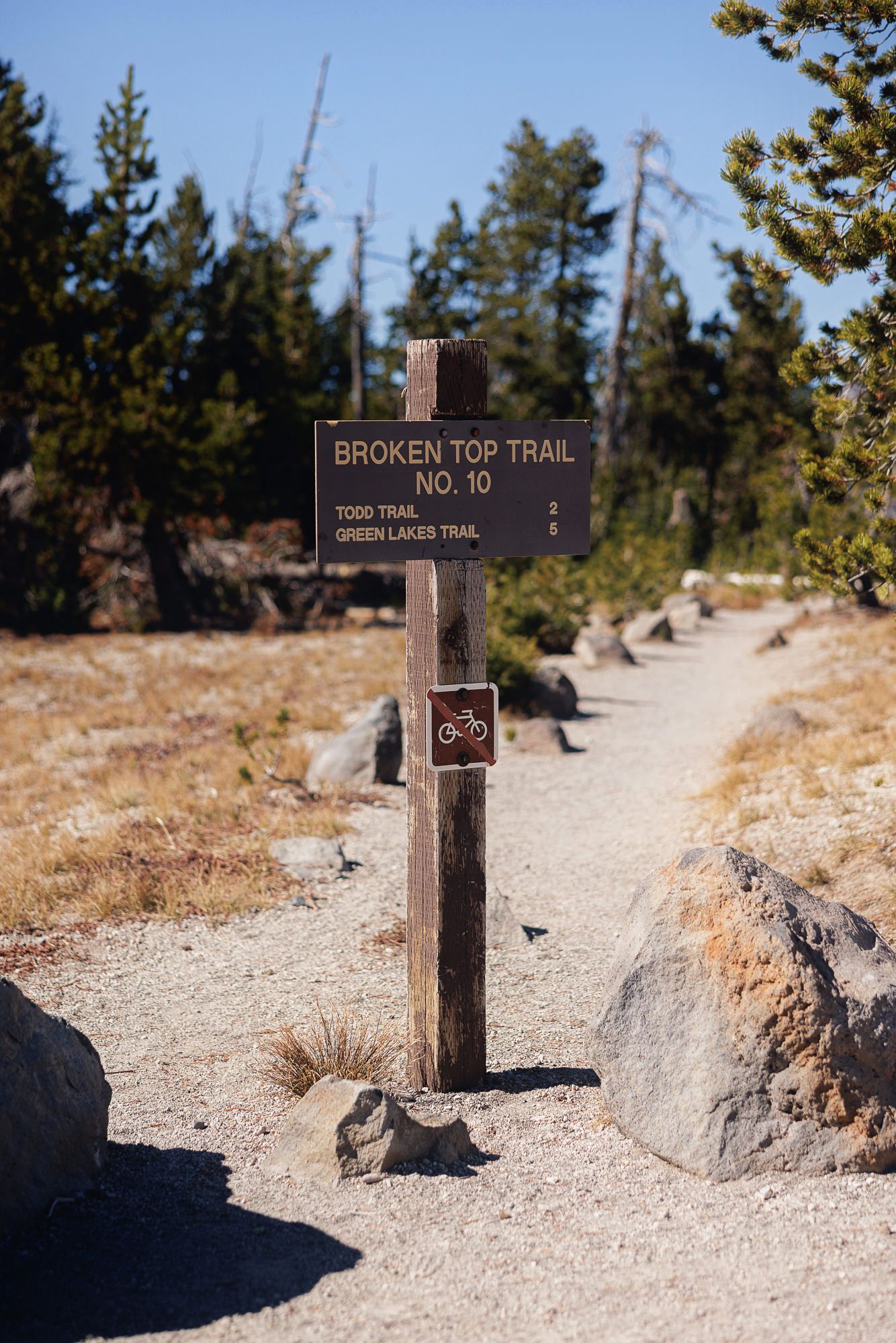 Wooden Broken Top Trail No. 10 sign with bike symbol beside a gravel path, boulders, and pine trees under a clear blue sky.