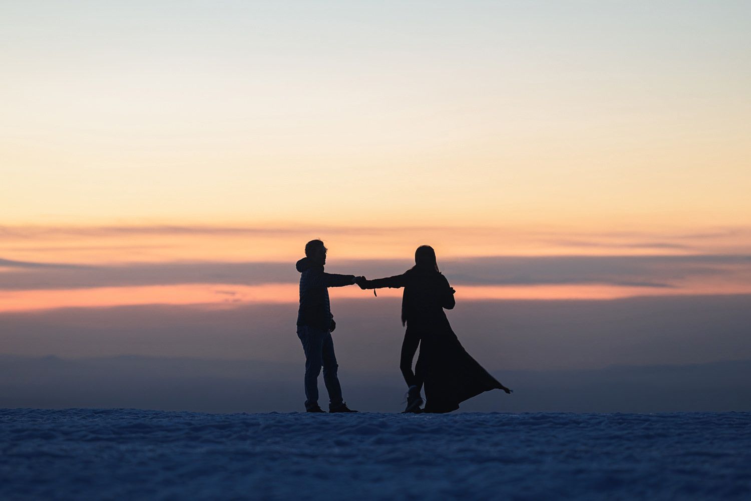 Two silhouettes share a romantic moment against a vibrant sunset sky over a snowy winter landscape.