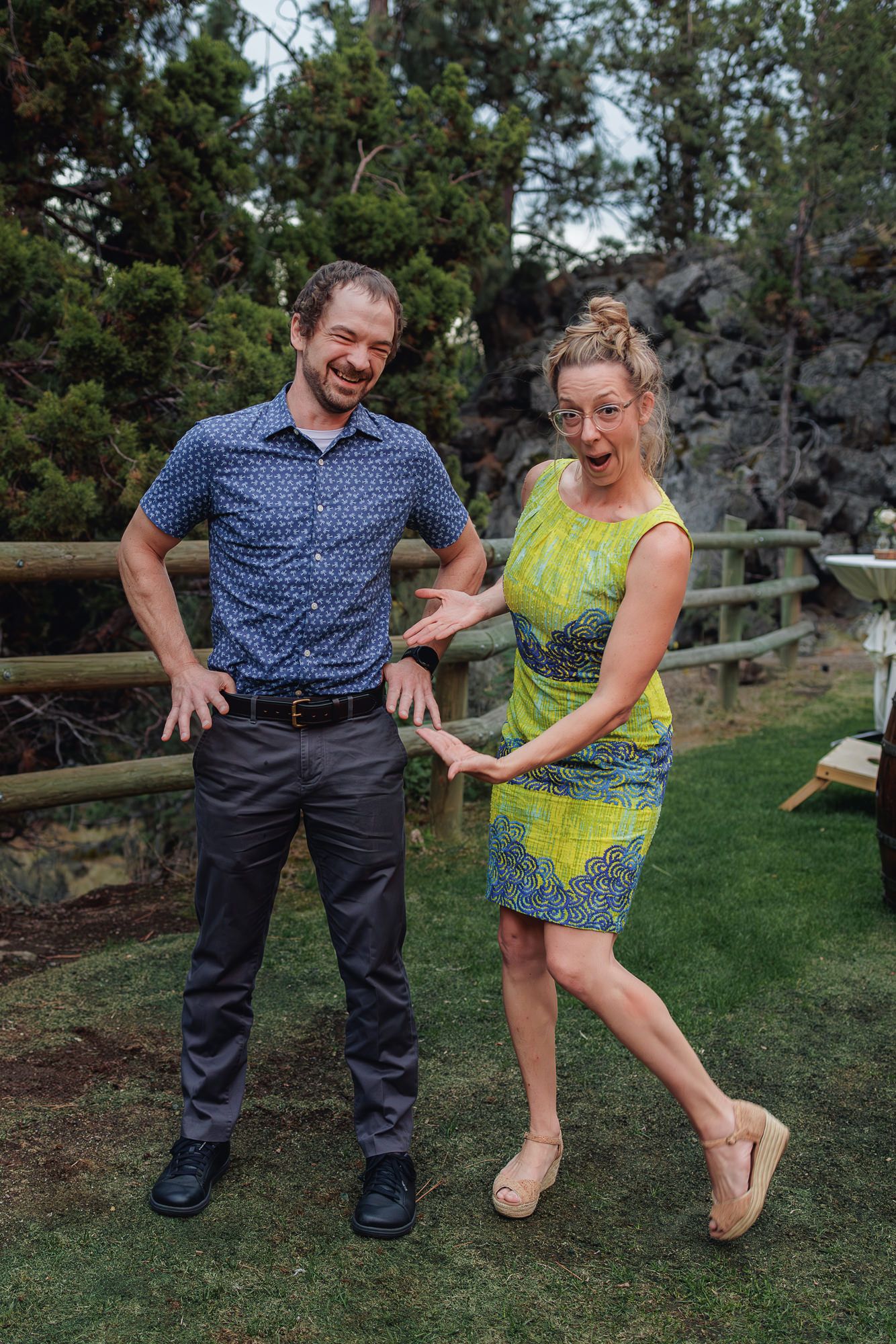A couple laughs together while posing playfully in a garden setting with trees and a wooden fence in the background.