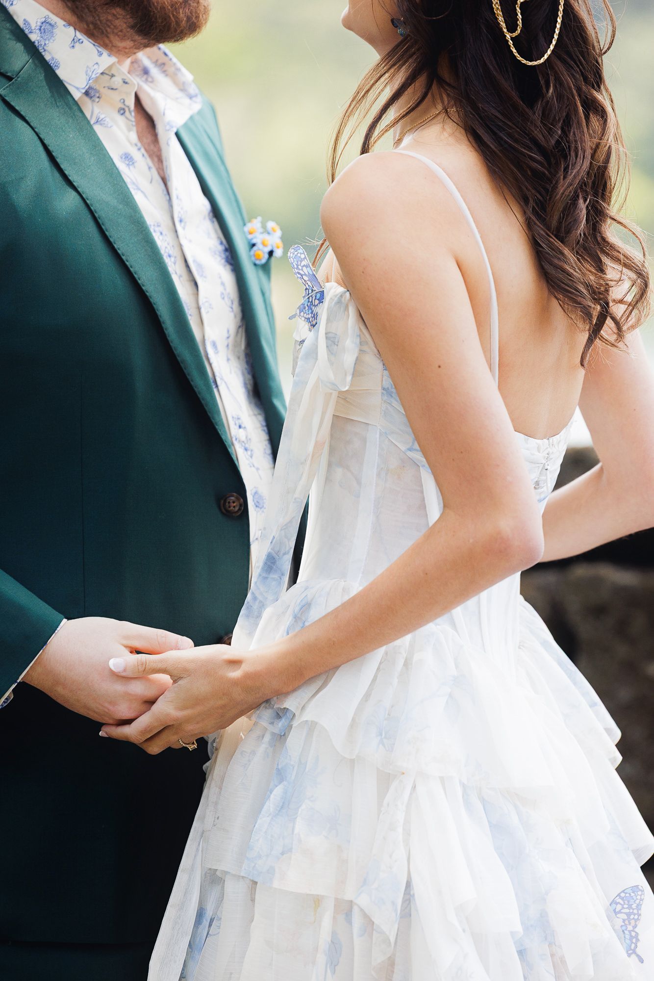 Couple holding hands at wedding, bride in blue floral tiered dress, groom in emerald green suit with floral shirt.