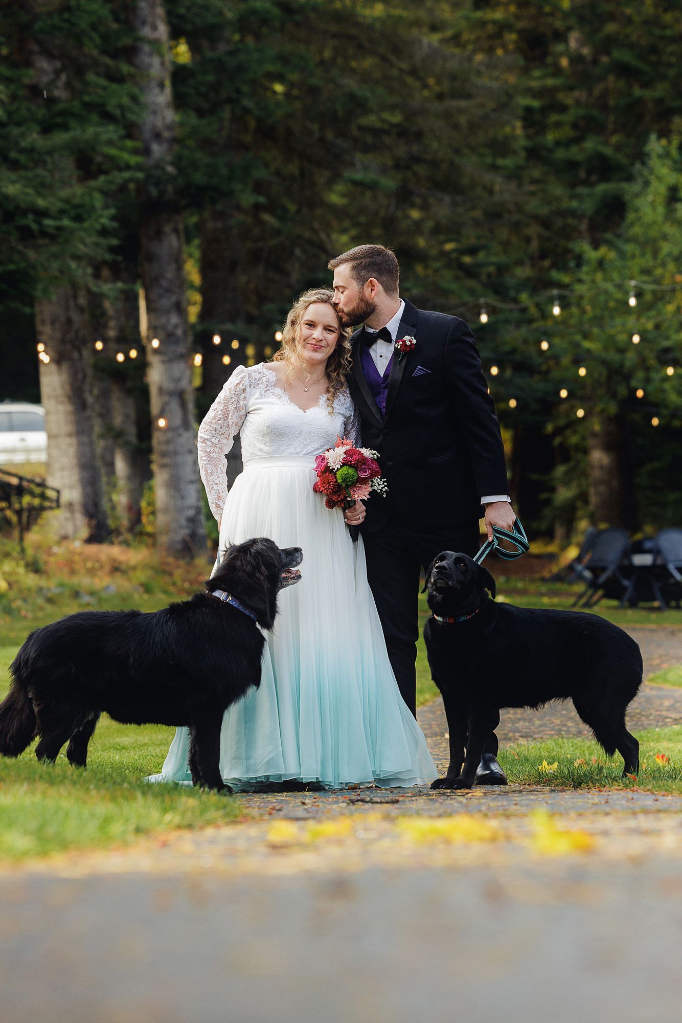 A couple in wedding attire sits on grass with their black dog during an outdoor photoshoot in a wooded setting.