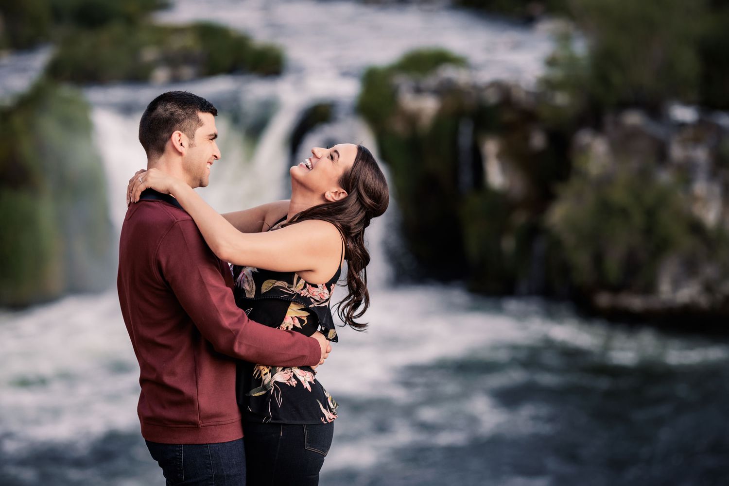 Couple embracing outdoors with a waterfall and natural scenery in the background.