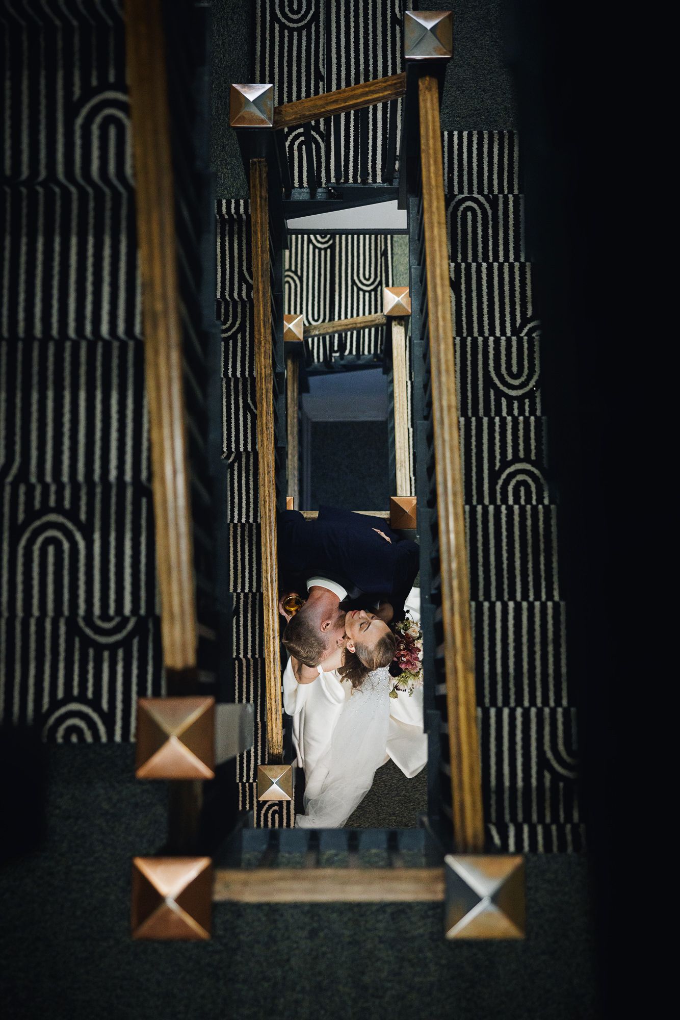 Couple sharing a romantic kiss on a staircase with elegant Art Deco patterned carpet and gold stud details.