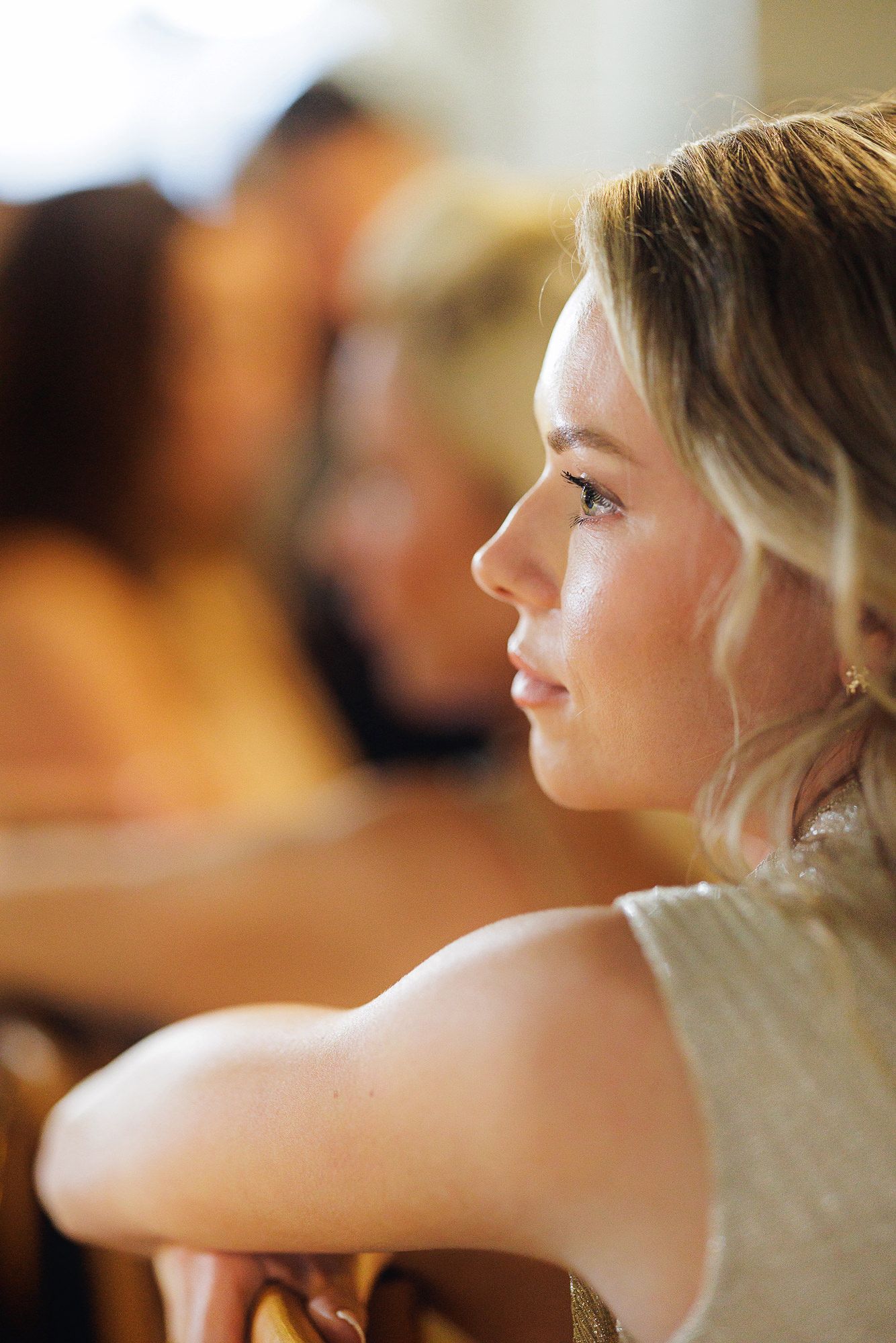Young blonde woman in profile view, gazing thoughtfully in a warmly lit indoor setting.