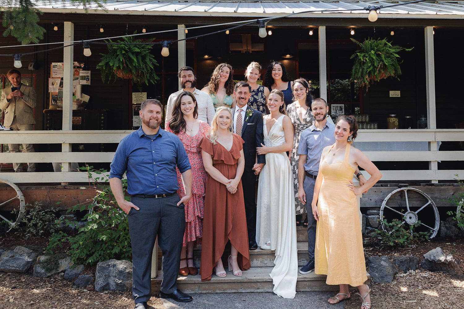 A group gathered on wooden porch steps outside of a rustic building with hanging plants and string lights.