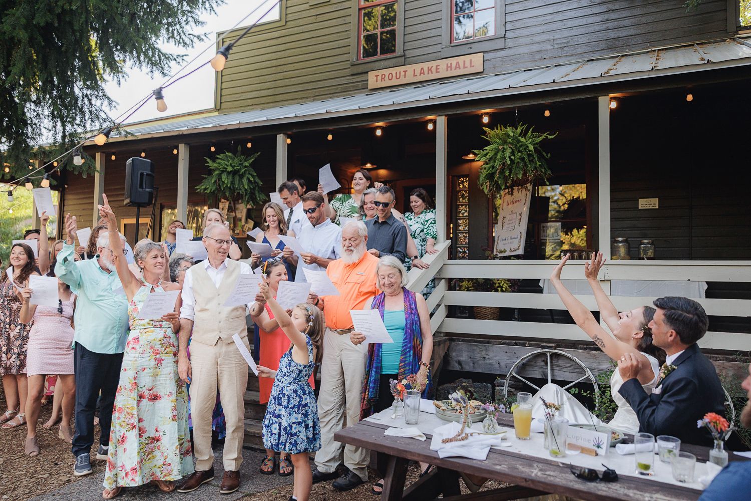 A large group gathers on the front porch of a gray house with hanging plants during an outdoor social event.