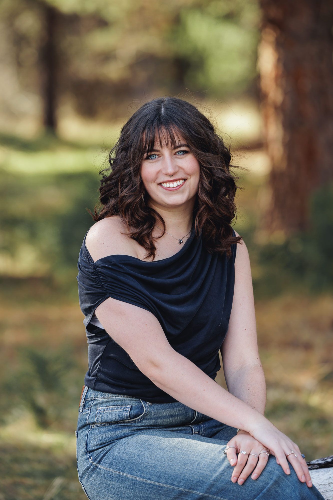 Smiling young woman with dark curly hair wearing a black top and jeans, sitting outdoors in a forest setting.