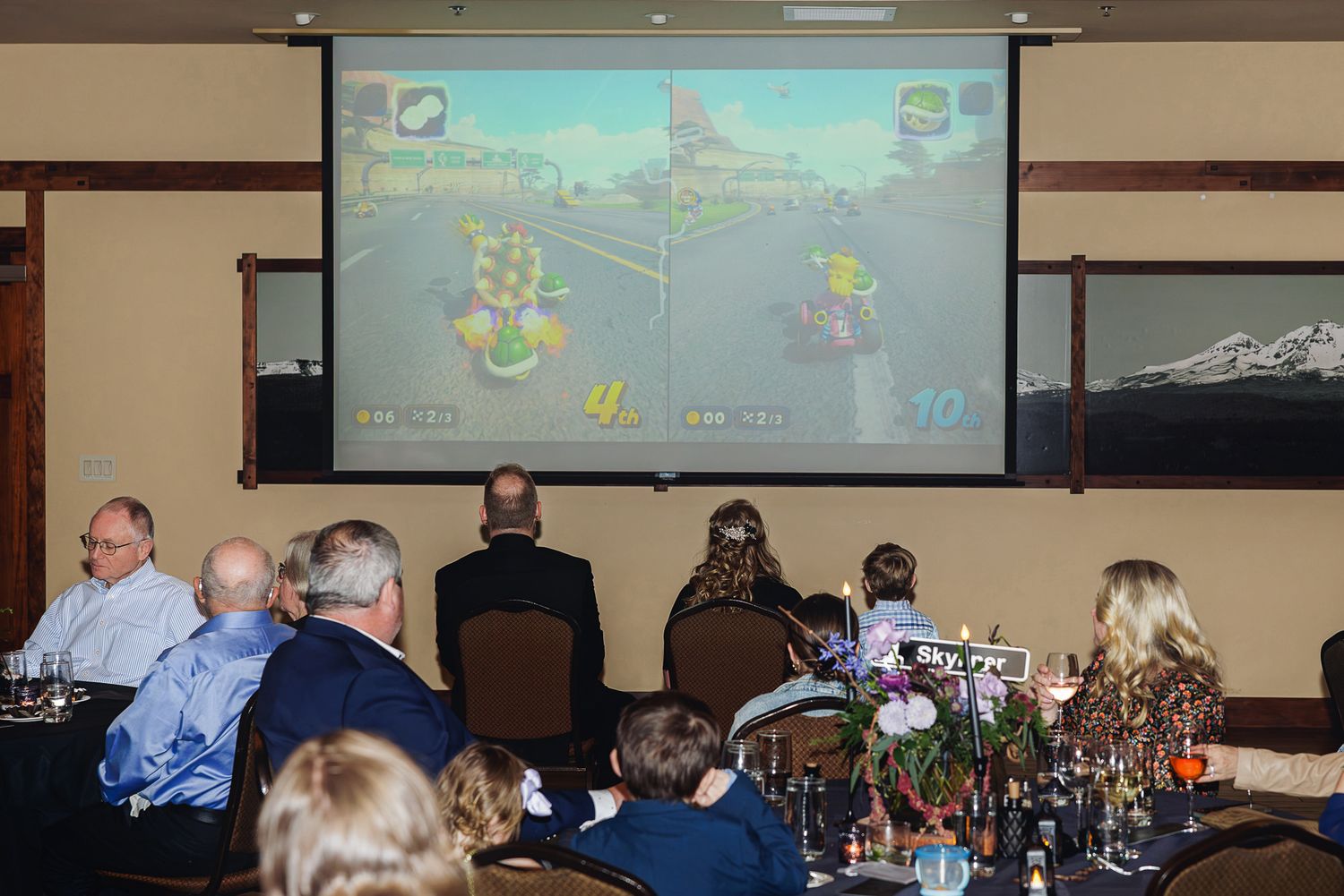 Group of people watching Mario Kart racing game projected on large screen during a formal dinner event.