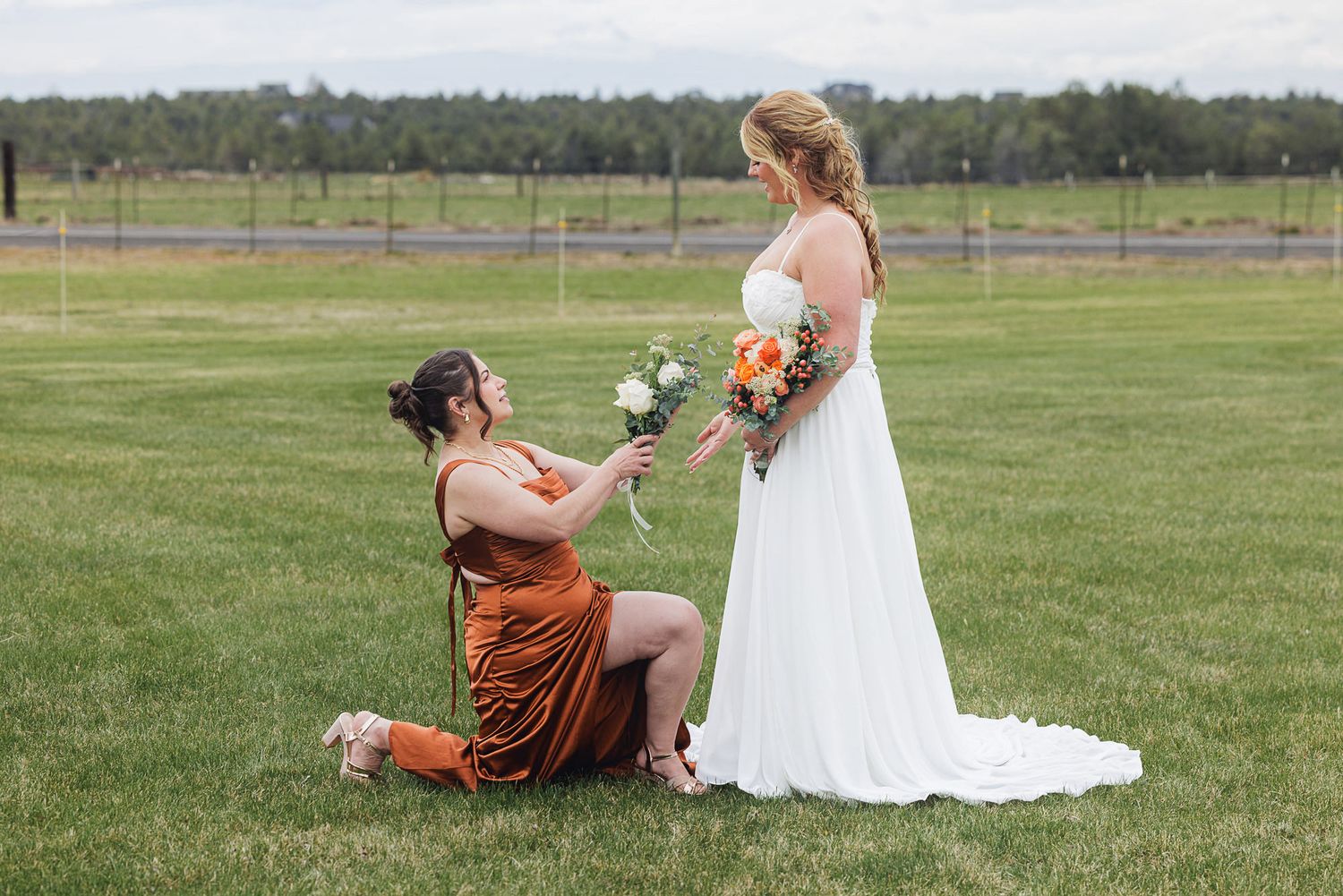 Person in orange dress kneeling to offer flowers to a bride in white gown on green lawn at outdoor wedding.