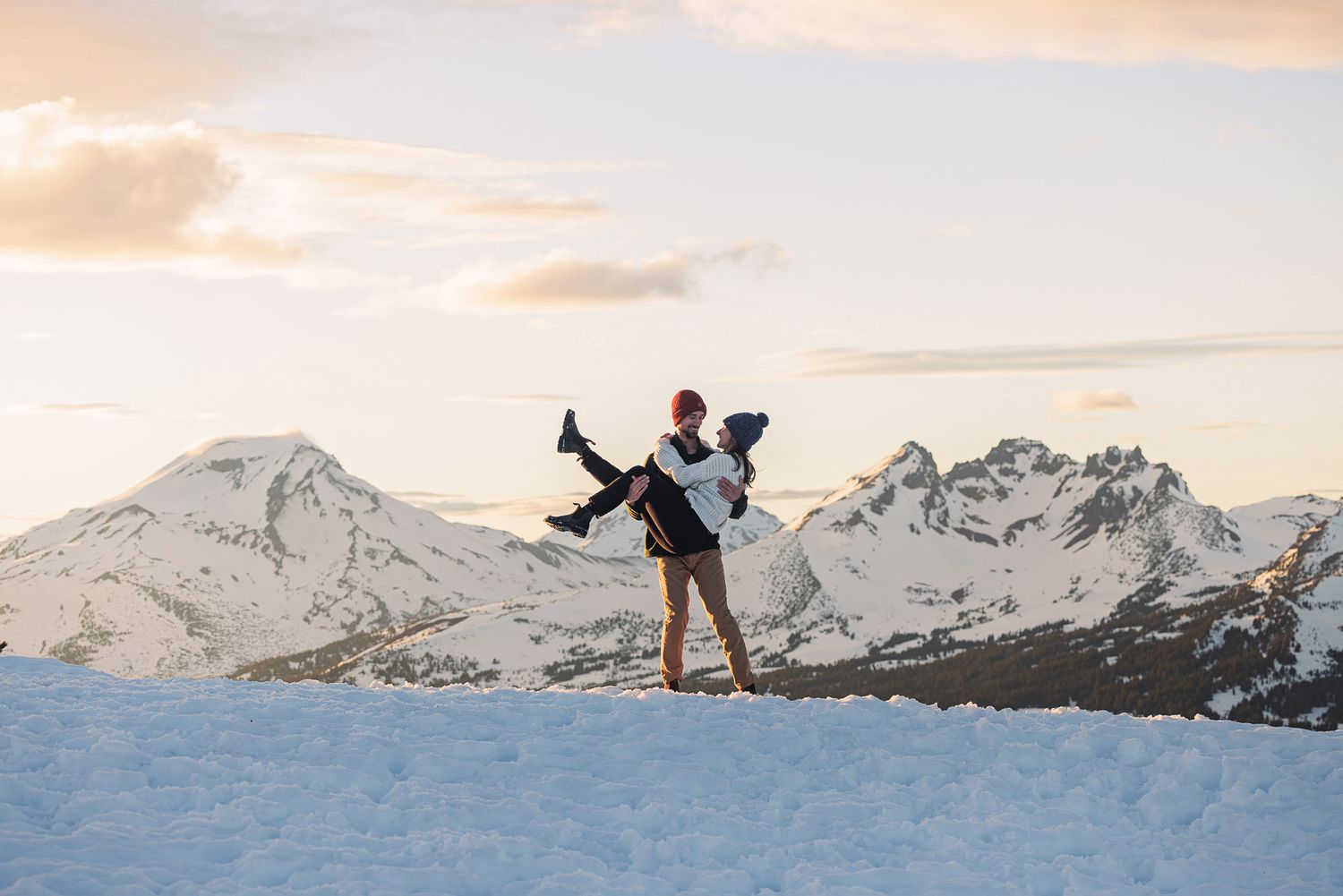 Couple embracing on a snowy mountain summit at sunset with snow-capped peaks in the background.