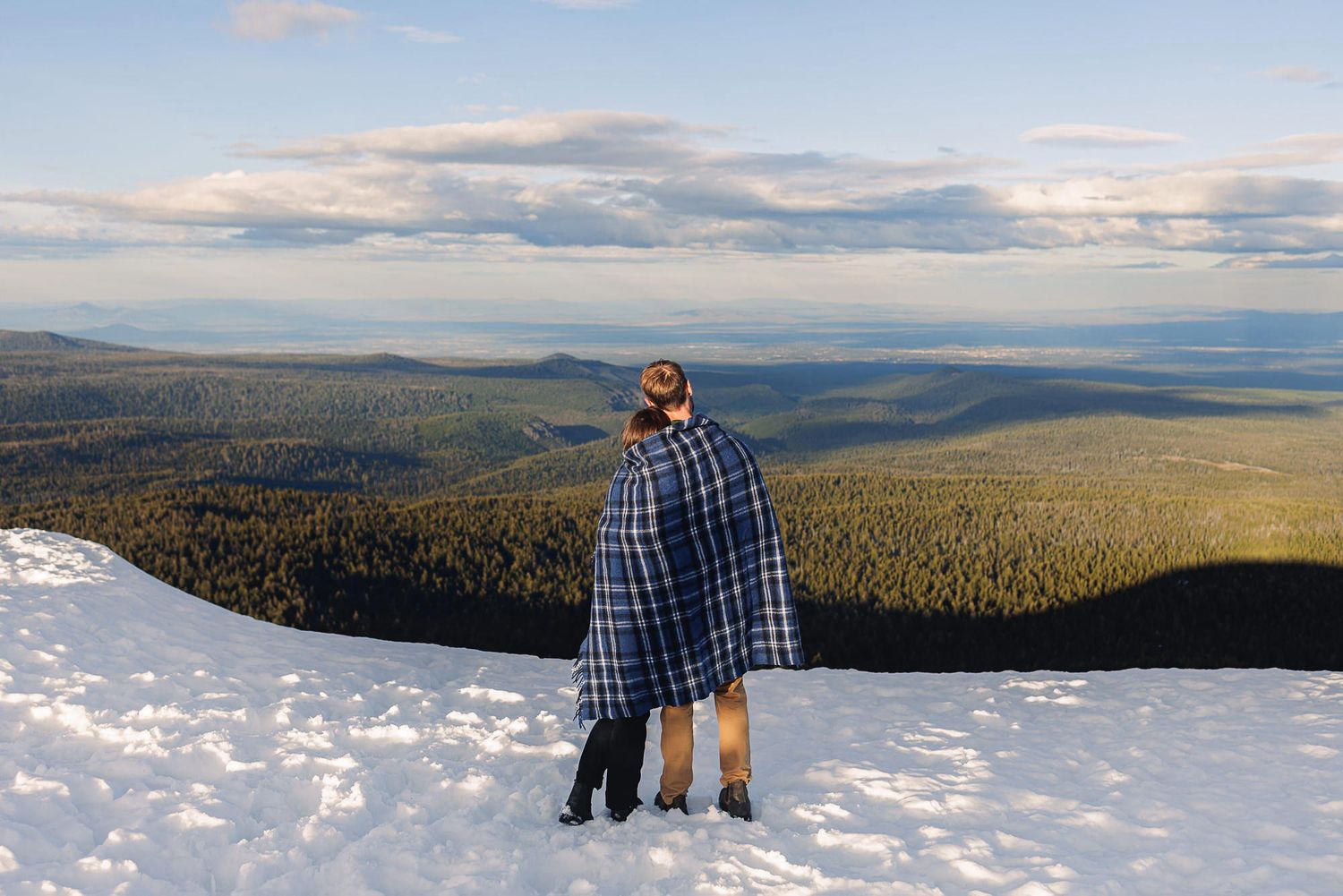 Couple wrapped in plaid blanket standing on snowy mountain peak overlooking vast forest landscape under cloudy sky.