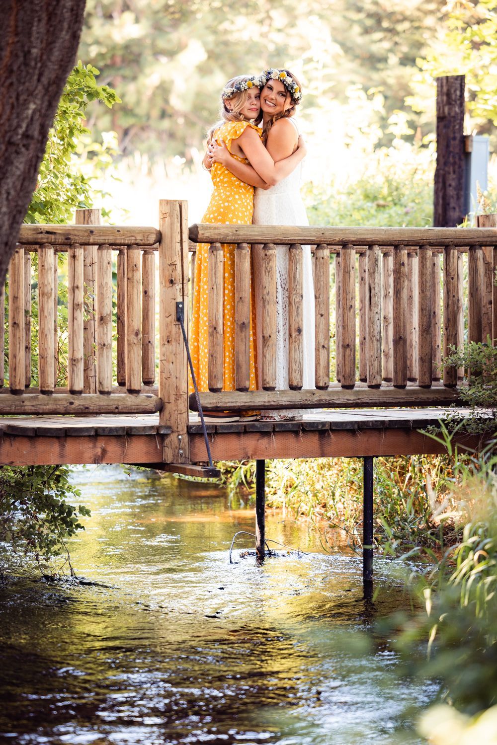 Two people embrace on a rustic wooden bridge over a peaceful stream in a sunlit forest setting.