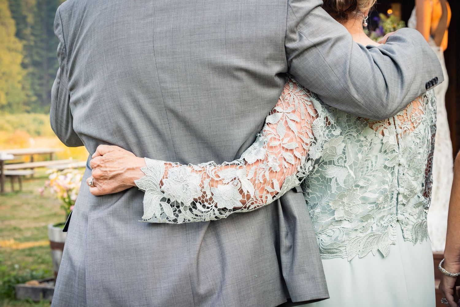 A close-up of a couple embracing, showing the bride's lace sleeve detail against the groom's gray suit.