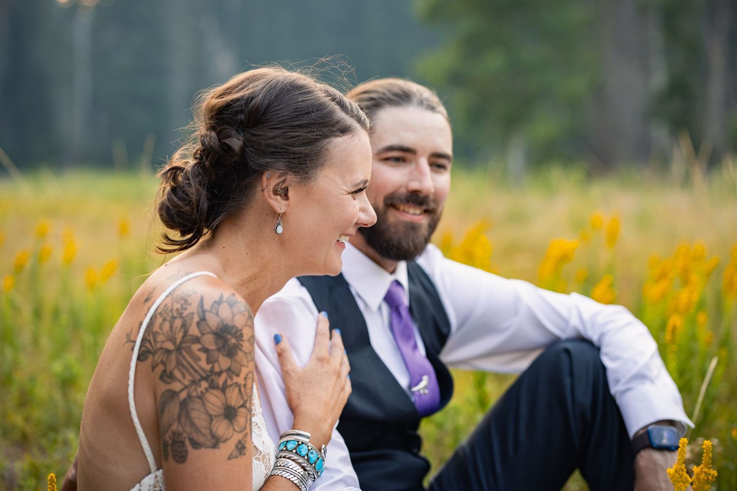 A happy couple sits together laughing in a meadow with bright yellow wildflowers in the background.