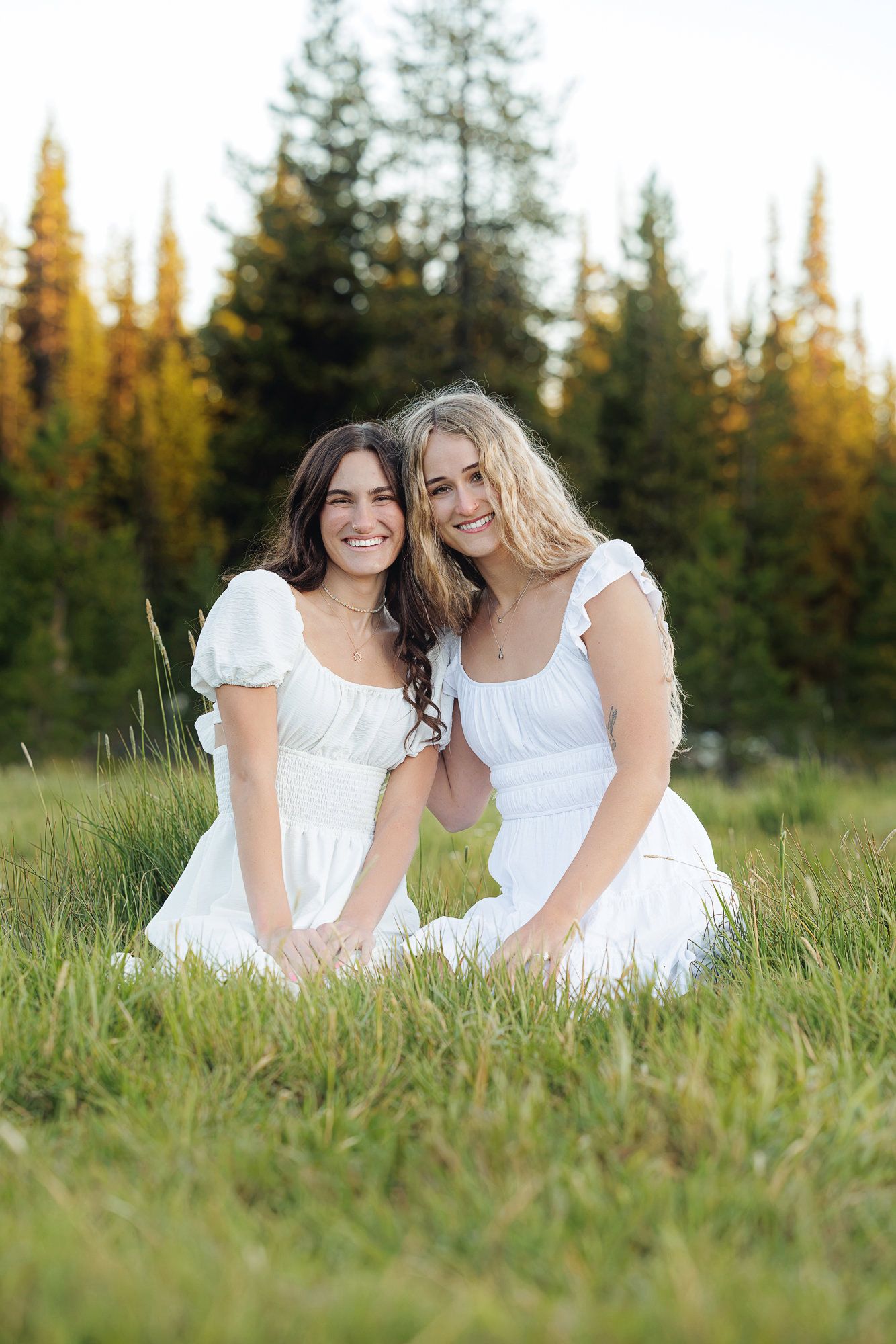 Two young women in white dresses sitting in a grassy meadow surrounded by pine trees, smiling warmly.