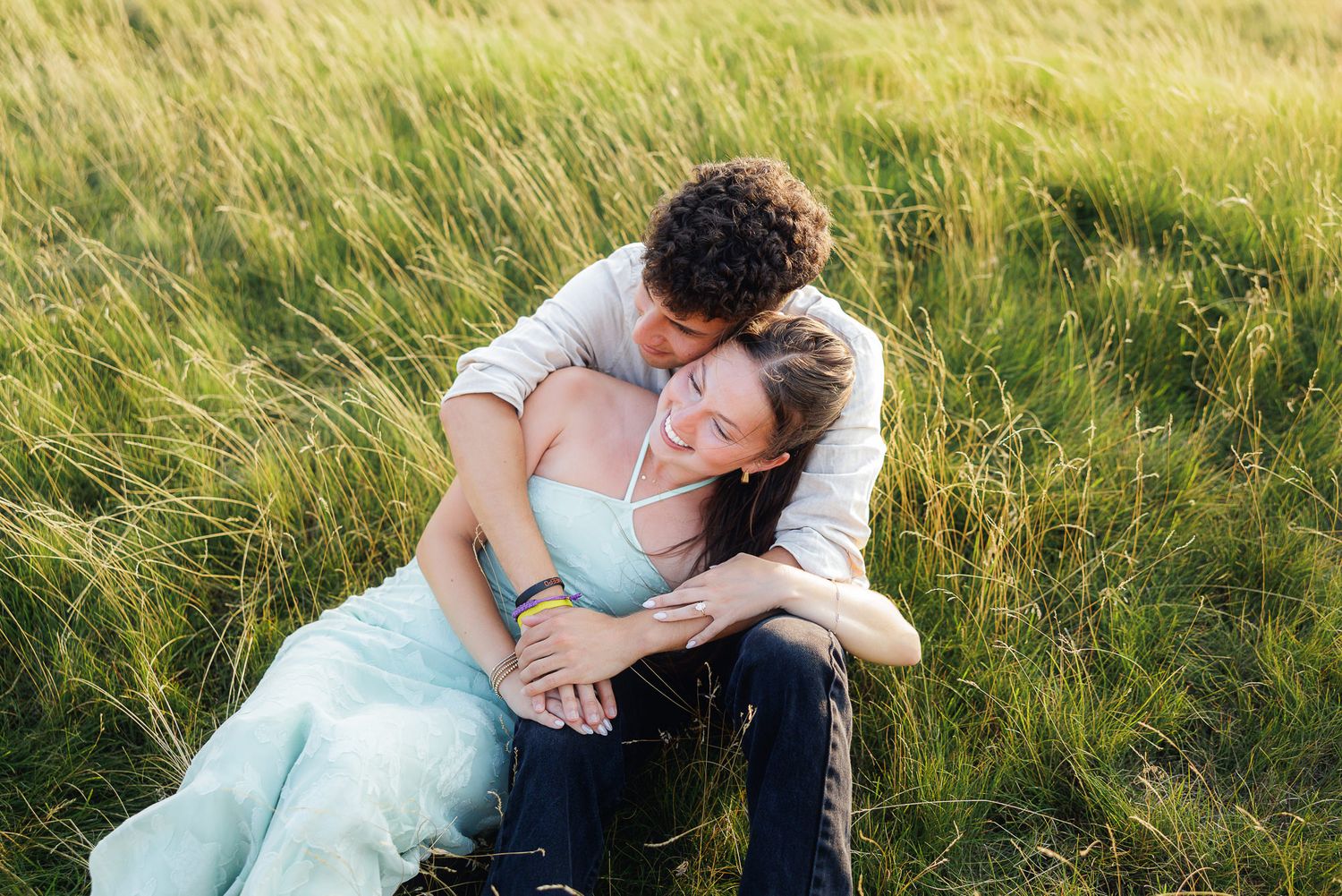 Couple sitting in tall green grass field, man embracing woman in light blue dress, romantic outdoor portrait.
