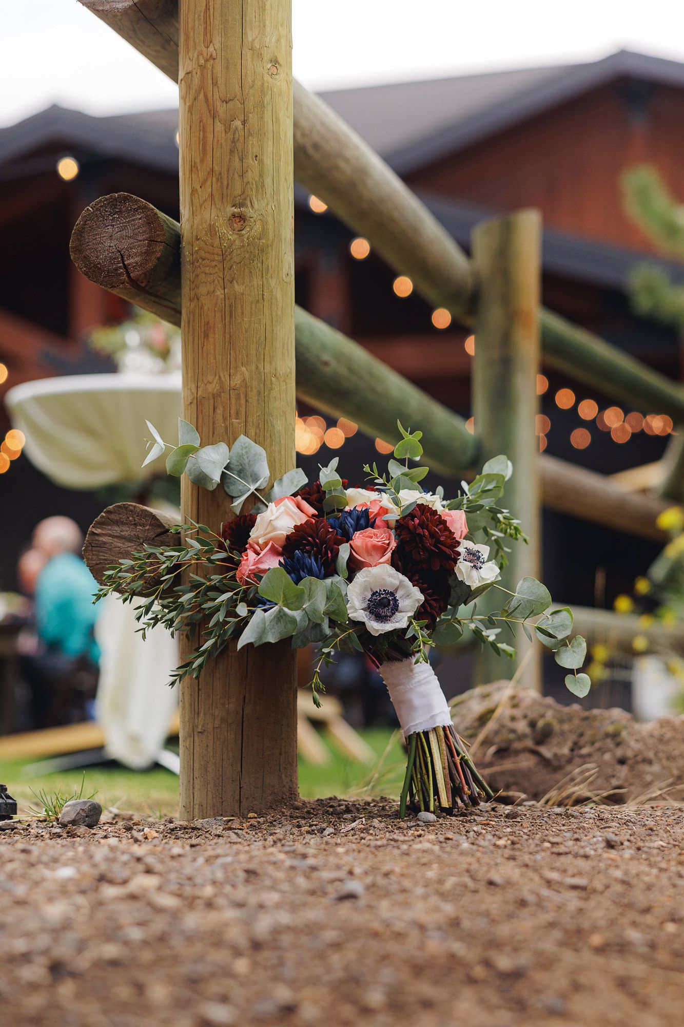 A rustic wedding bouquet with burgundy and blush flowers rests against a wooden post under string lights at an outdoor venue.