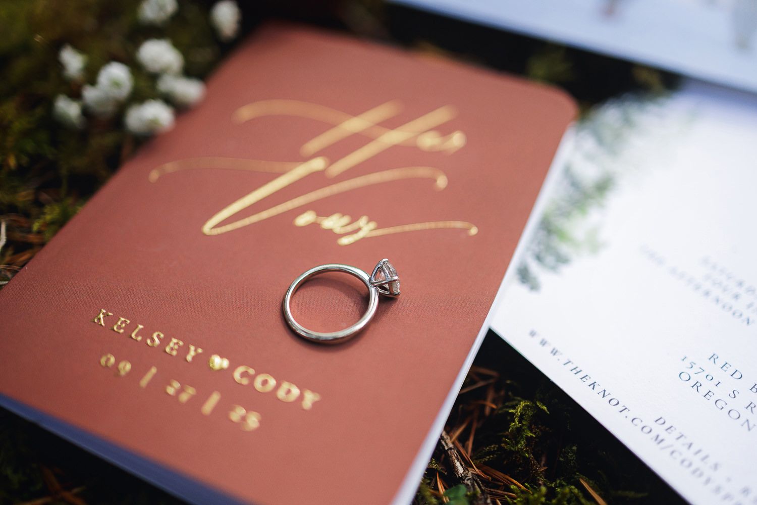 A diamond engagement ring rests on a terracotta-colored wedding invitation with gold lettering.