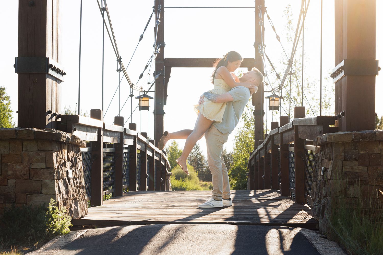 Couple sharing a romantic embrace on a wooden suspension bridge at golden hour with warm sunlight glowing behind them.
