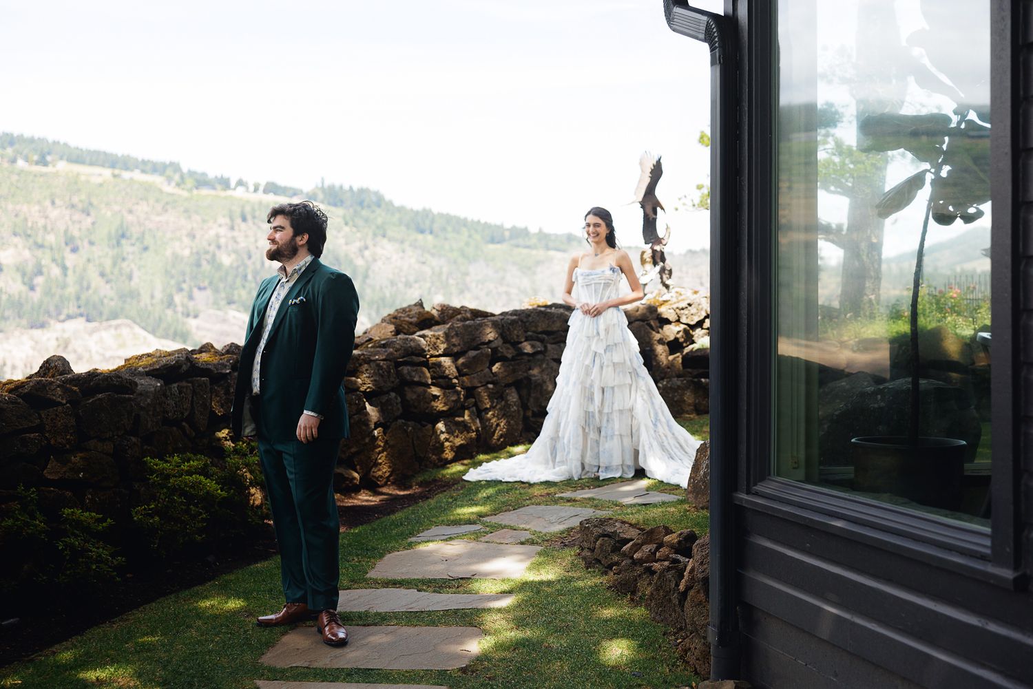 Bride in white gown and groom in teal suit stand apart on stone terrace with scenic mountain views.